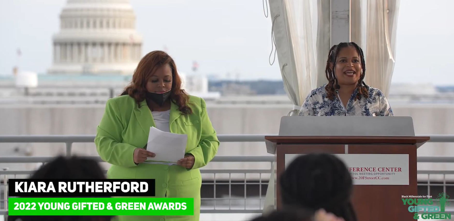 Two women standing at a podium during an outdoor event with the U.S. Capitol building in the background. One woman is reading from papers, and the other is speaking at the microphone. There is a banner with event details and a logo in the bottom right corner.