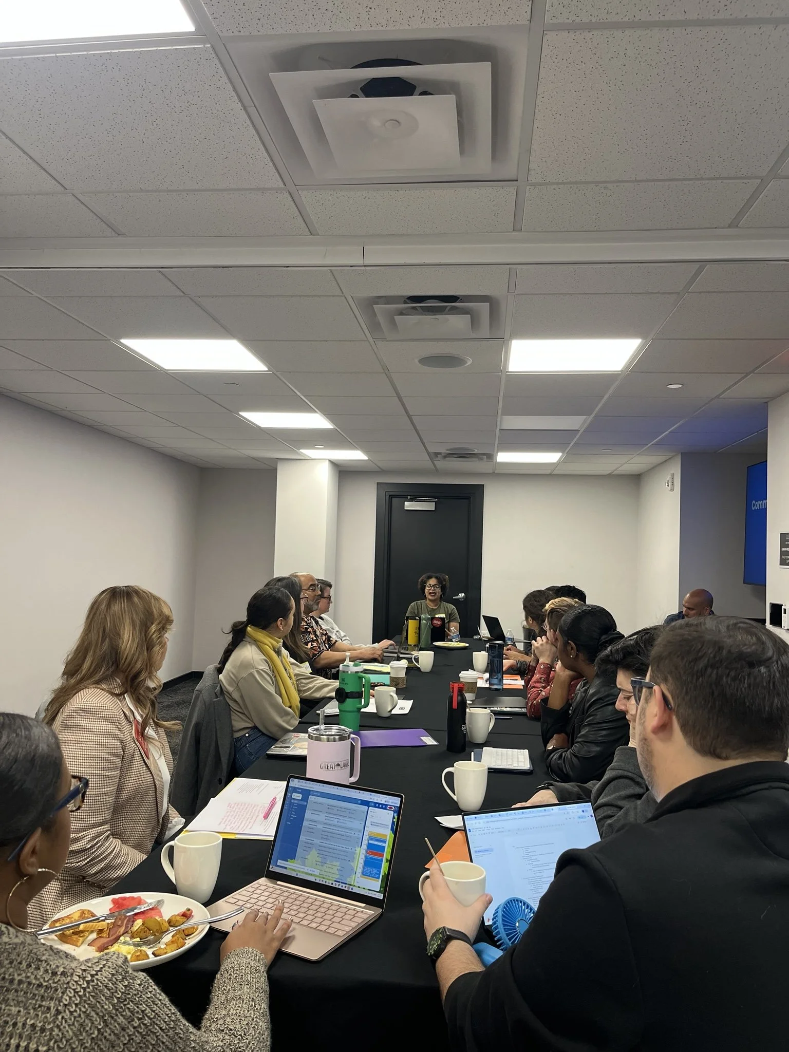 A diverse group of people sitting around a long conference table in a meeting room, with a woman speaking at the head of the table and laptops, notebooks, and coffee cups on the table.