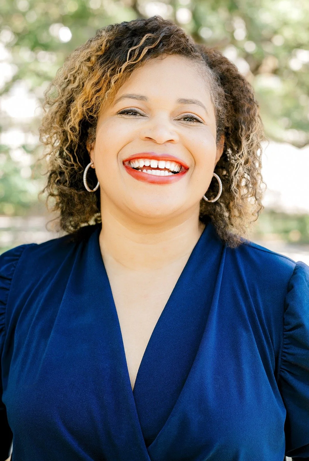 A woman smiling outdoors, wearing a blue top and hoop earrings, with curly hair and a background of greenery.