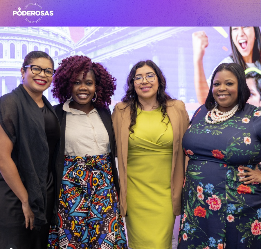 Four women standing together at a conference or summit, smiling for the camera. The background displays the words 'Poderosas Summit 2024' and words indicating a legislative theme, with an illustrated image of Capitol building.