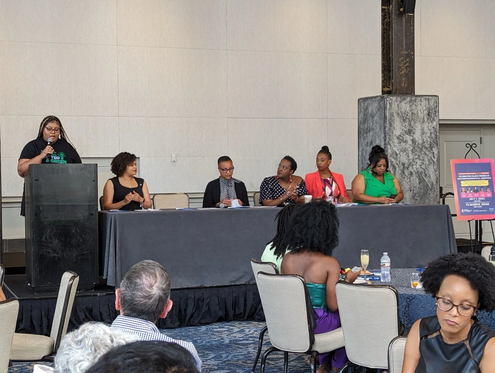 A panel of six women sitting at a long table on a stage, with one woman standing at a podium speaking into a microphone, at an indoor conference event. Audience members are seated in front.