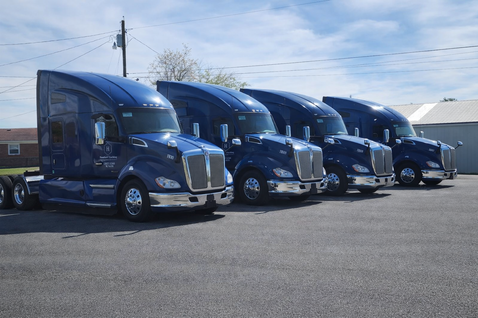 Four blue semi-trucks parked side by side on a paved lot with a building, electrical pole, and power lines in the background.