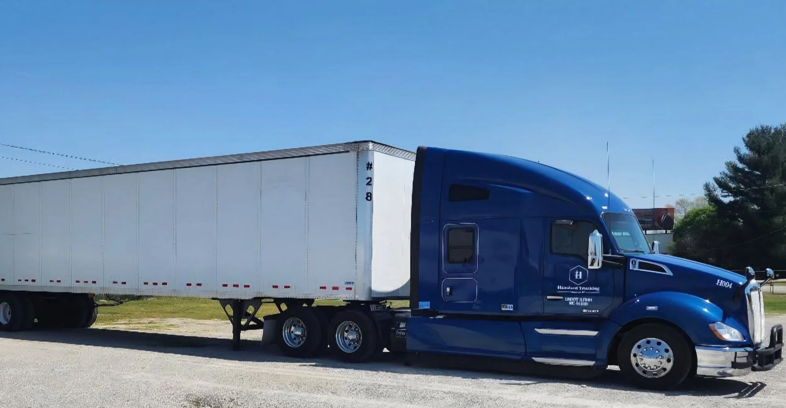 A blue semi-truck with a white trailer parked on the side of a road under a clear blue sky.