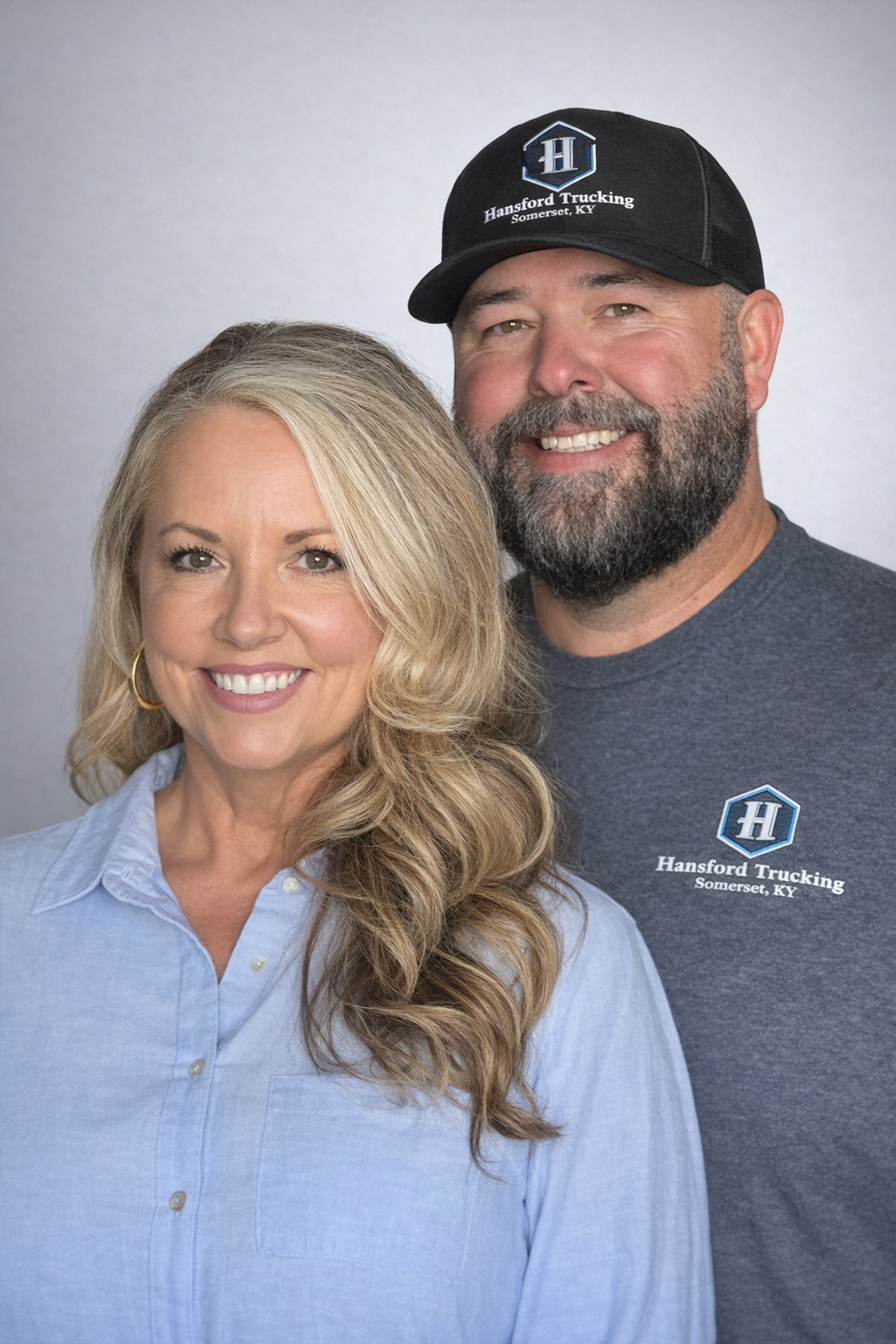 A smiling woman with blonde hair wearing a light blue button-down shirt and gold hoop earrings, standing next to a smiling man with a beard, wearing a black baseball cap and a dark gray t-shirt with a logo for Hansford Trucking, Somerset, KY.
