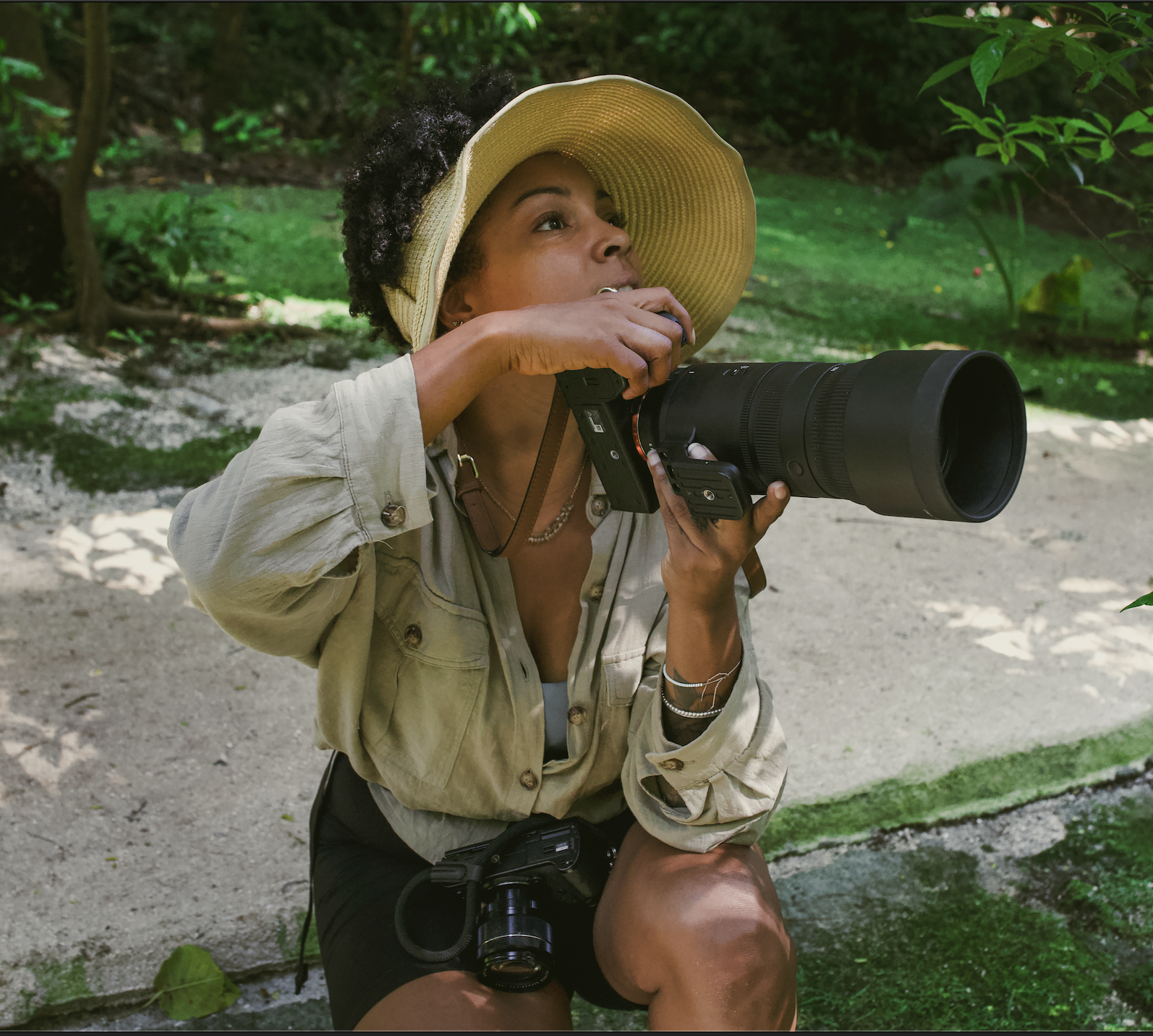A woman wearing a large yellow sun hat and a beige jacket is holding a camera with a large telephoto lens, aiming it upward in a green outdoor setting.
