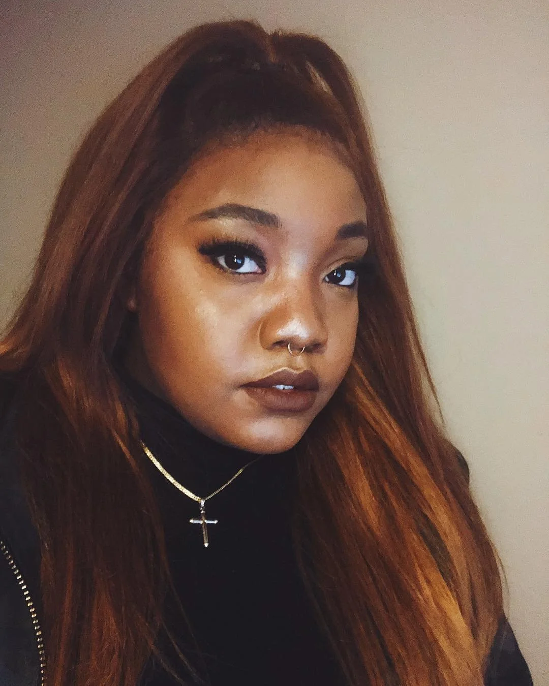 A young woman with long reddish-brown hair, wearing a black top, gold necklace with a cross pendant, and a septum piercing, looking directly into the camera against a neutral background.