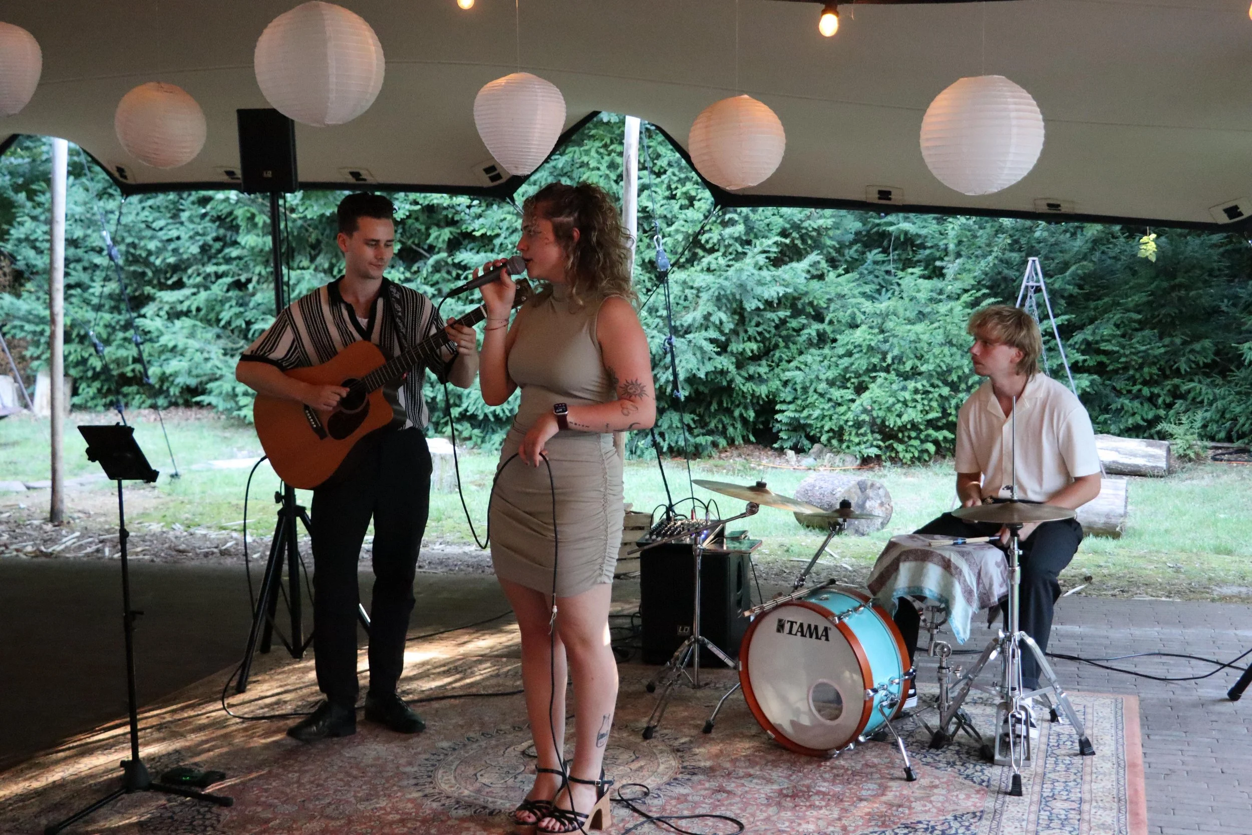 A band of three members performing outdoors under a tent, with paper lanterns hanging from the ceiling and surrounded by green trees.