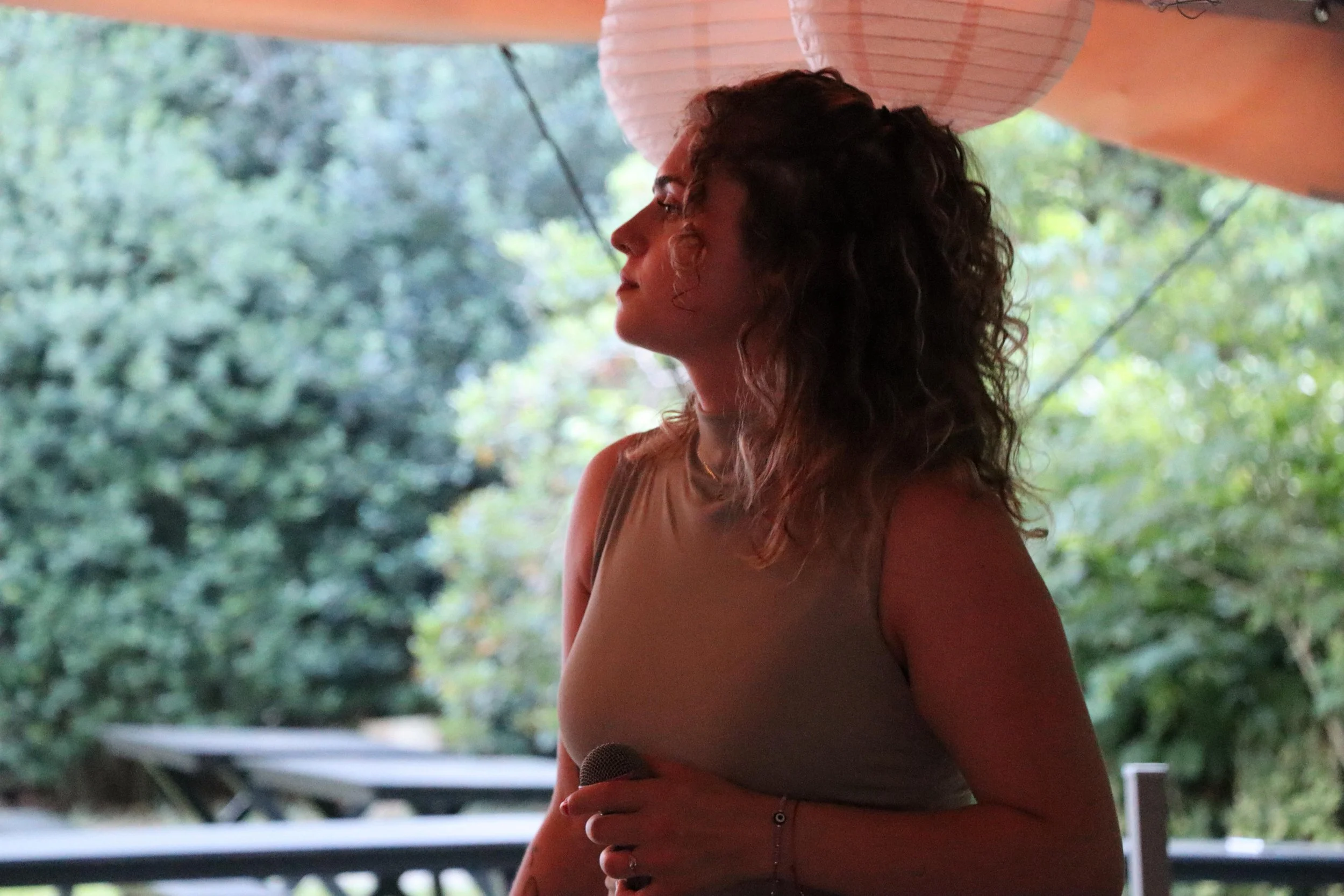 A woman with curly hair standing outdoors under a pink paper lantern, with green trees in the background.