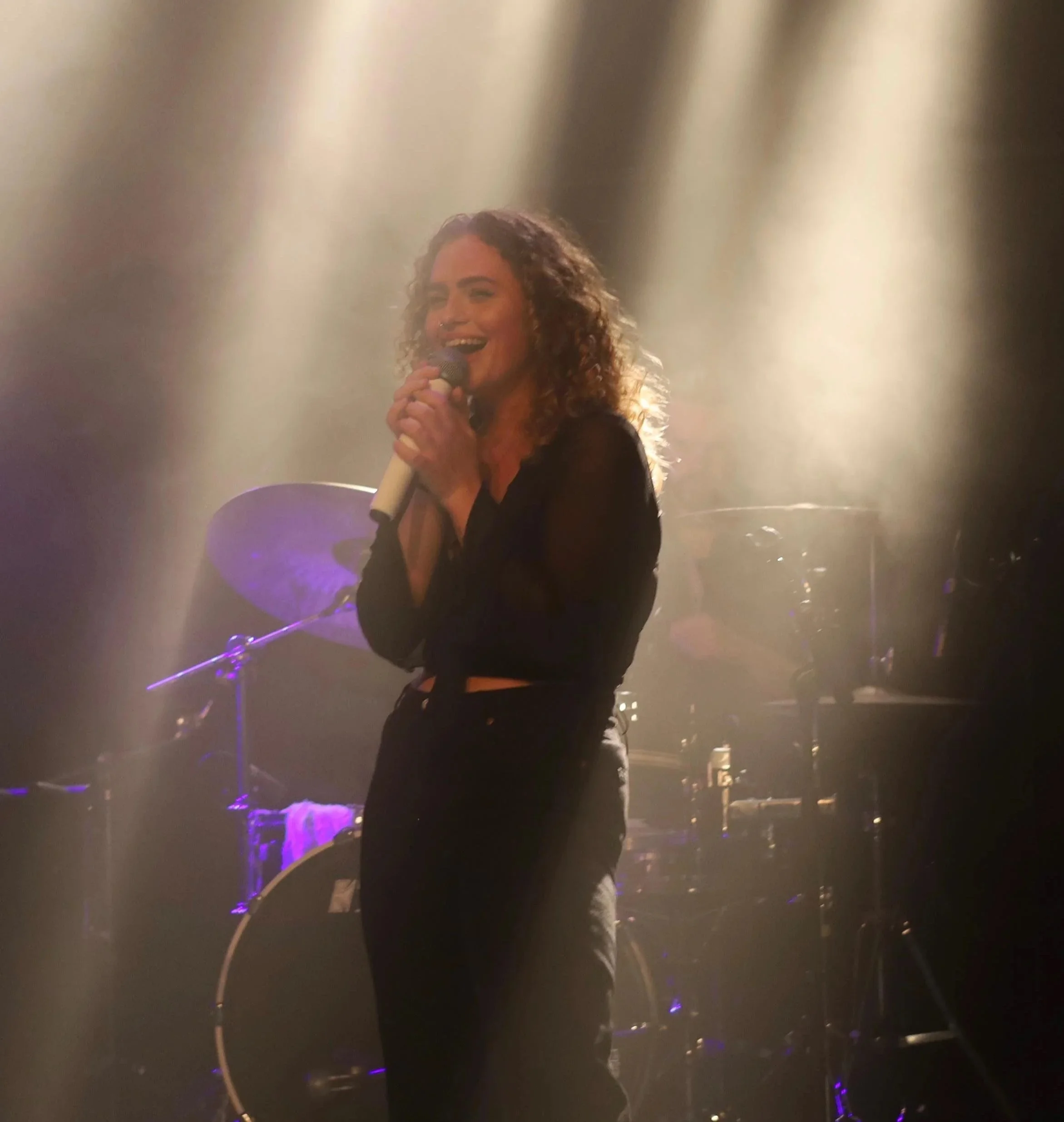 A woman with curly hair singing into a microphone on stage, illuminated by stage lights with purple and golden hues, with musical instruments visible in the background.
