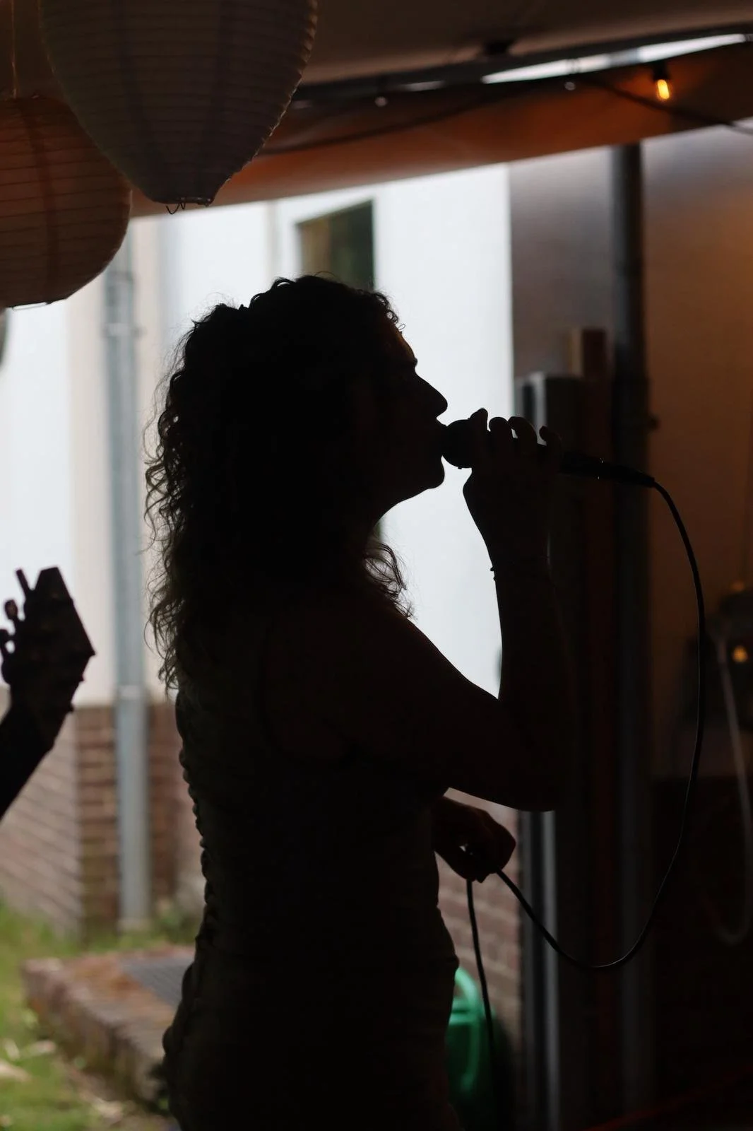 Silhouette of a woman singing into a microphone indoors with paper lanterns hanging above, during daytime.