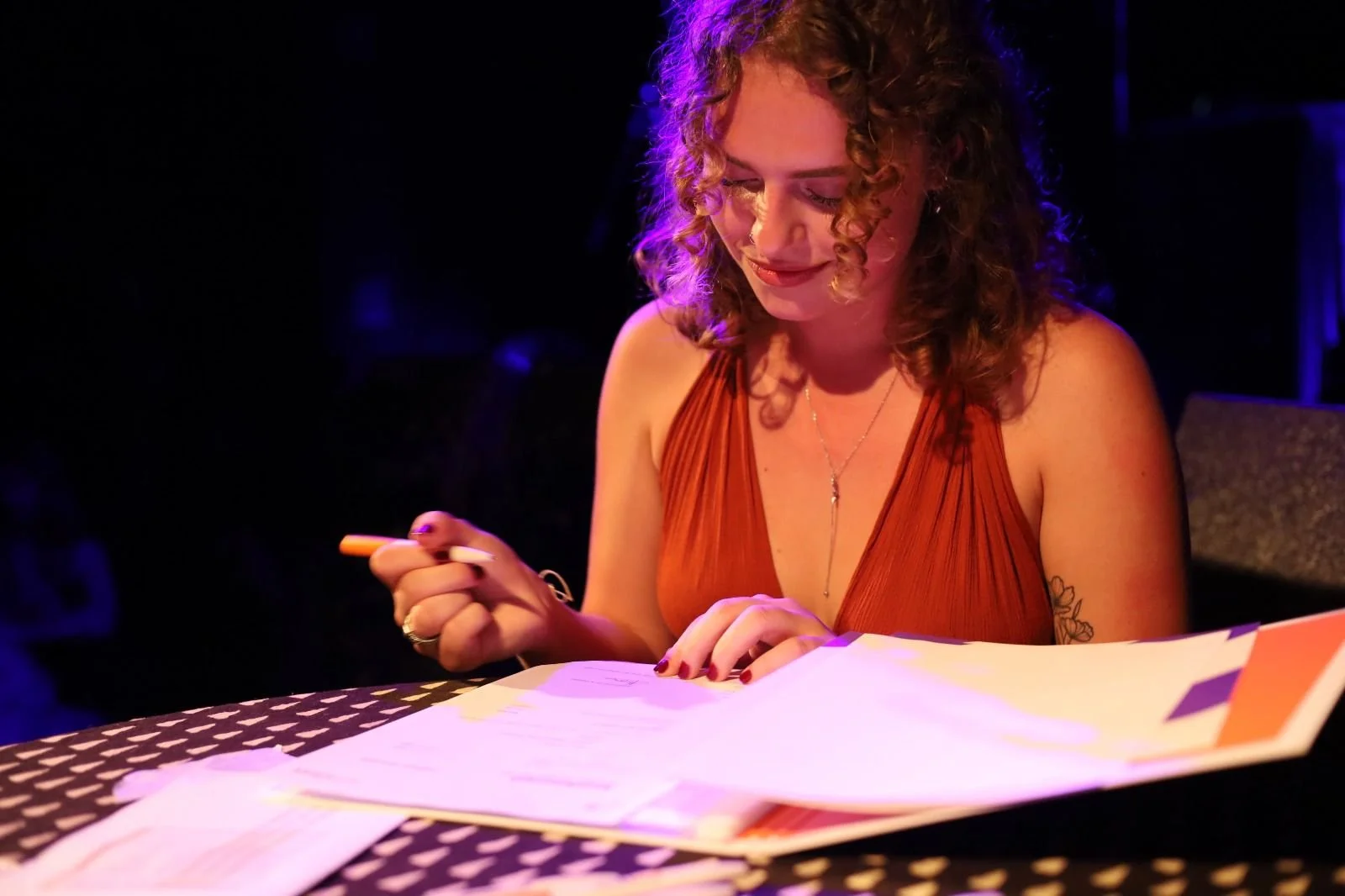 A woman with curly hair, wearing an orange sleeveless top, sits at a table with papers and a pen, looking down and smiling gently.