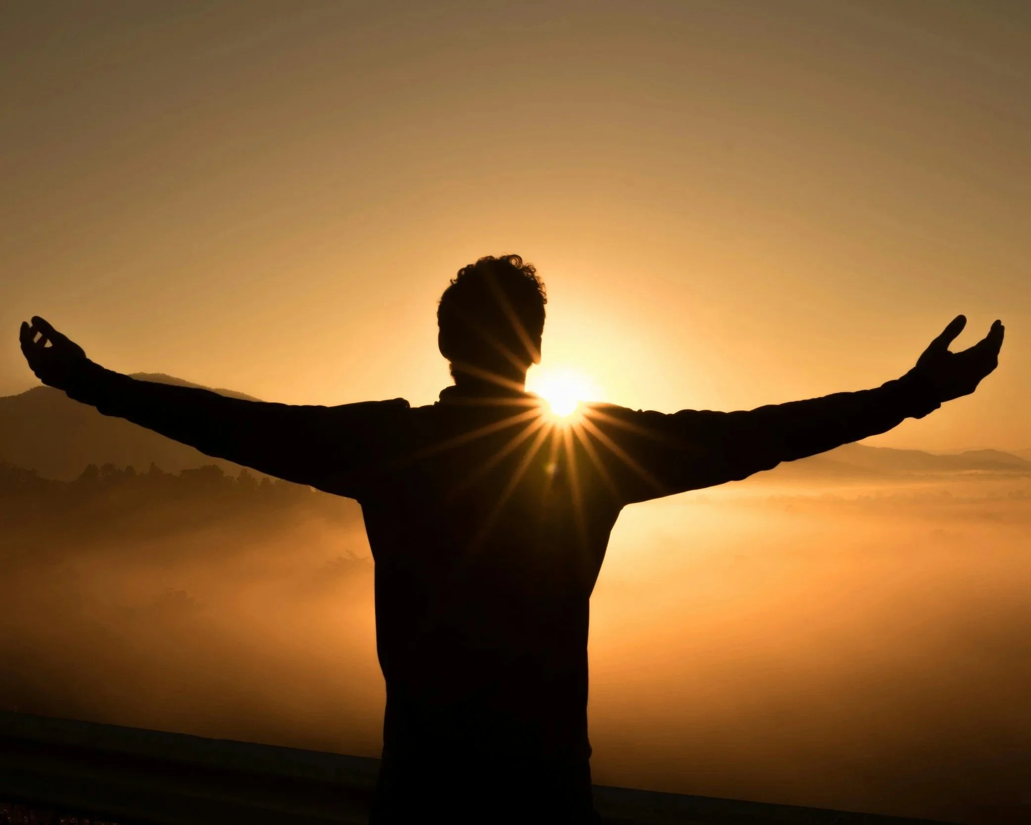 Silhouette of a person with arms outstretched in front of a sunset or sunrise with mountains and water in the background.