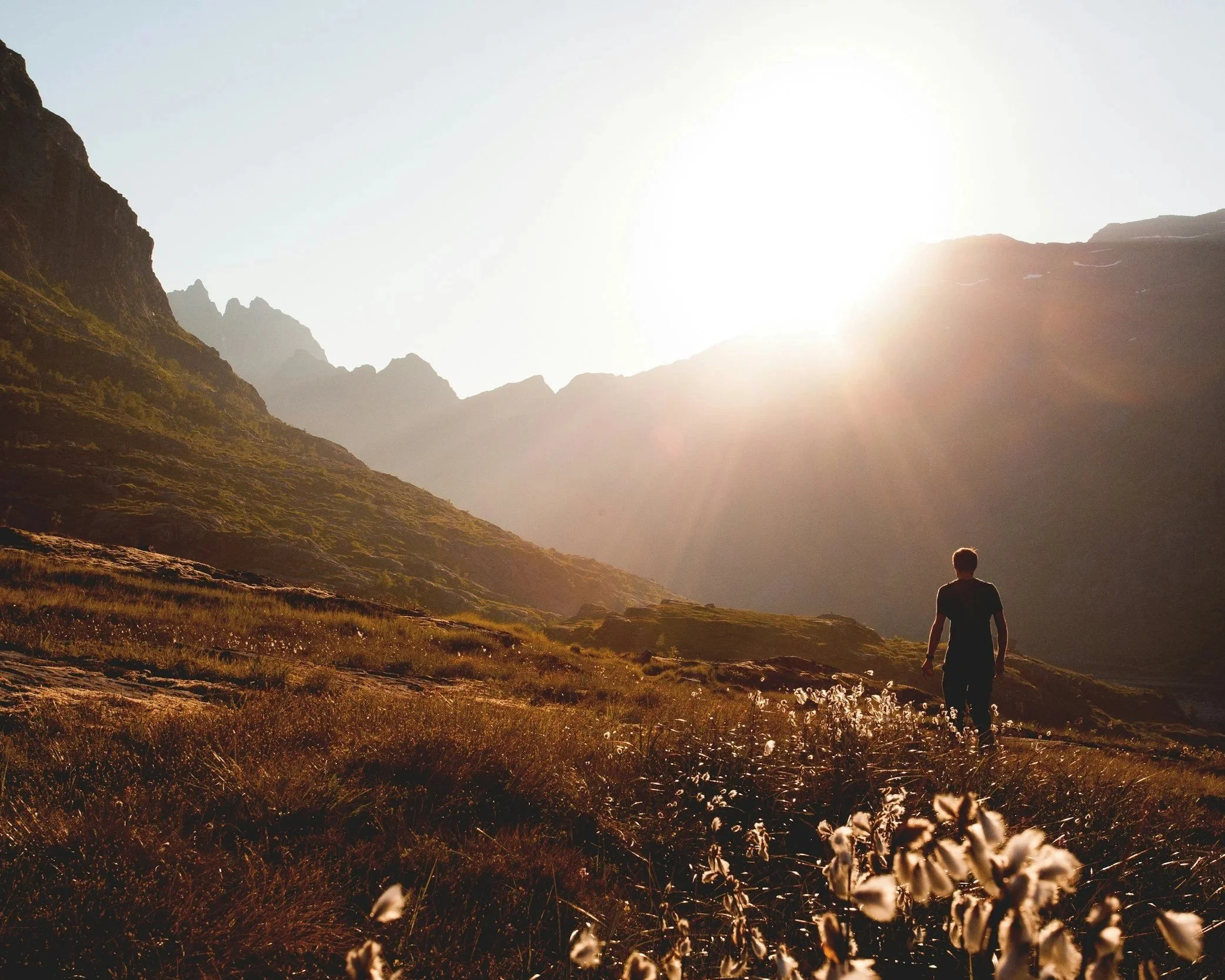 A person walking in a scenic mountainous landscape with the sun shining brightly, illuminating the hills and a valley with grass and wildflowers.