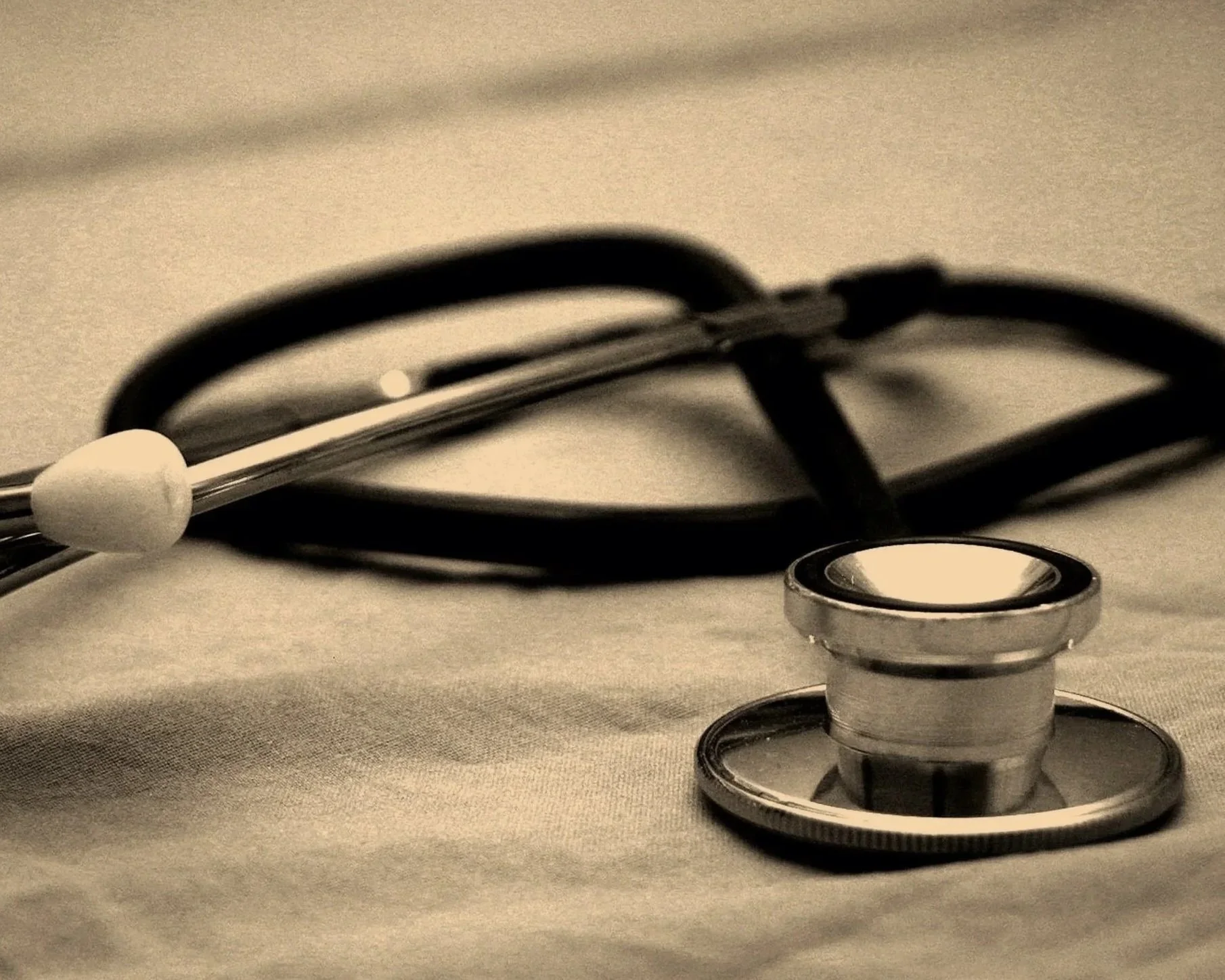 A stethoscope with black tubing and metal chest piece laid on a surface along with a silver medical ring, with a warm beige background.