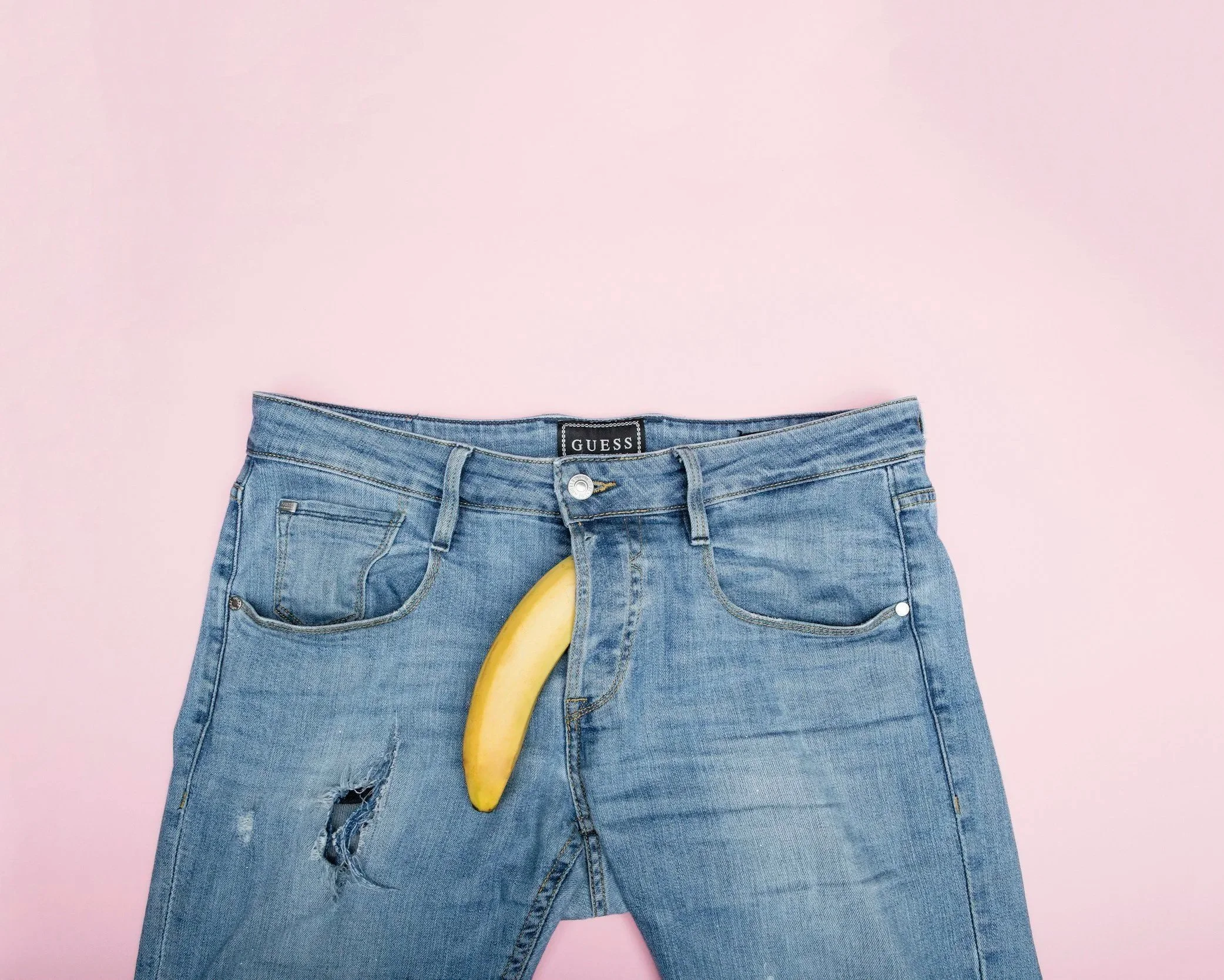 Pair of blue jeans with a ripped knee, with a yellow banana placed in the front pocket, against a pink background.