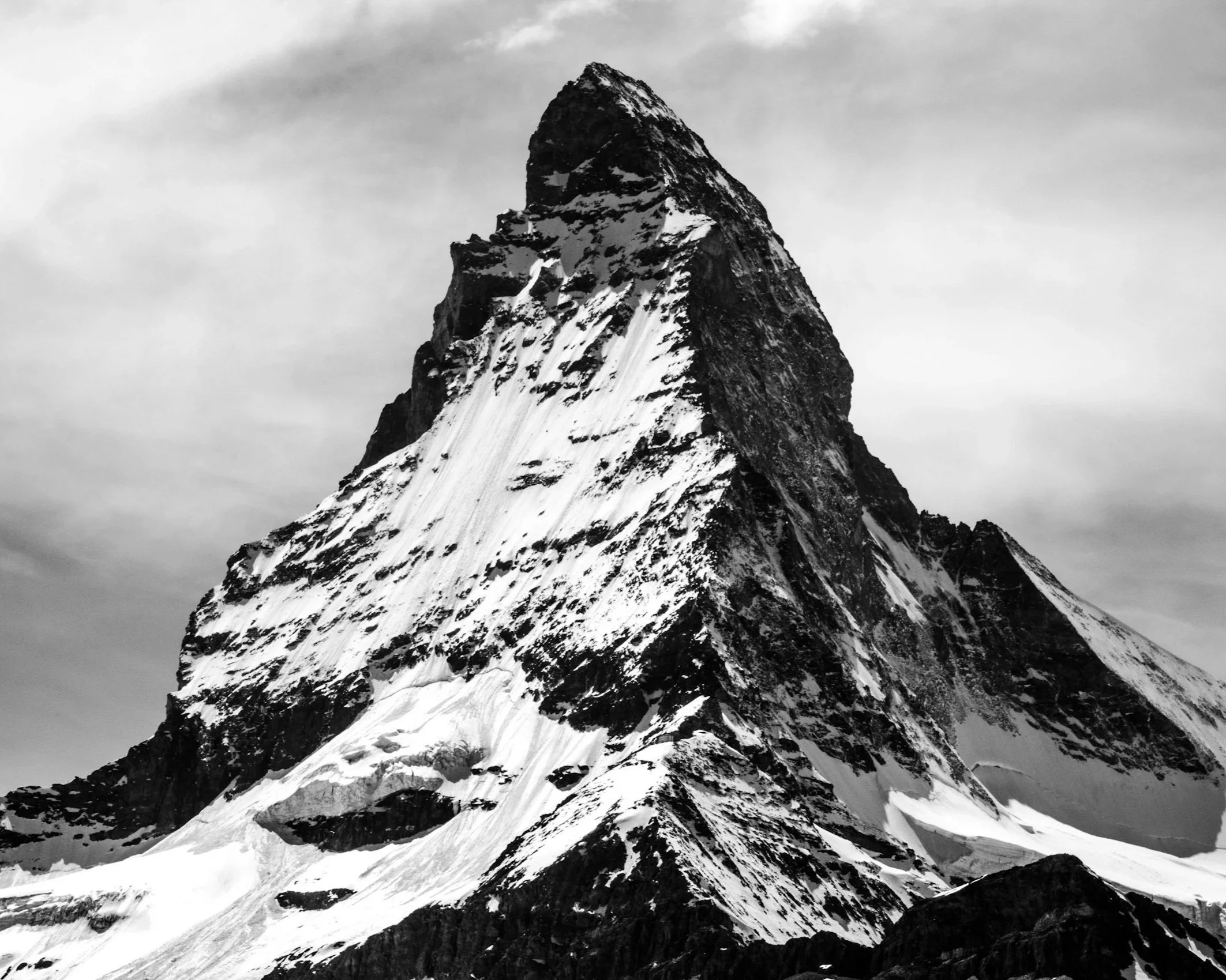 A black and white photograph of a prominent mountain peak covered with snow and ice, with a cloudy sky in the background.