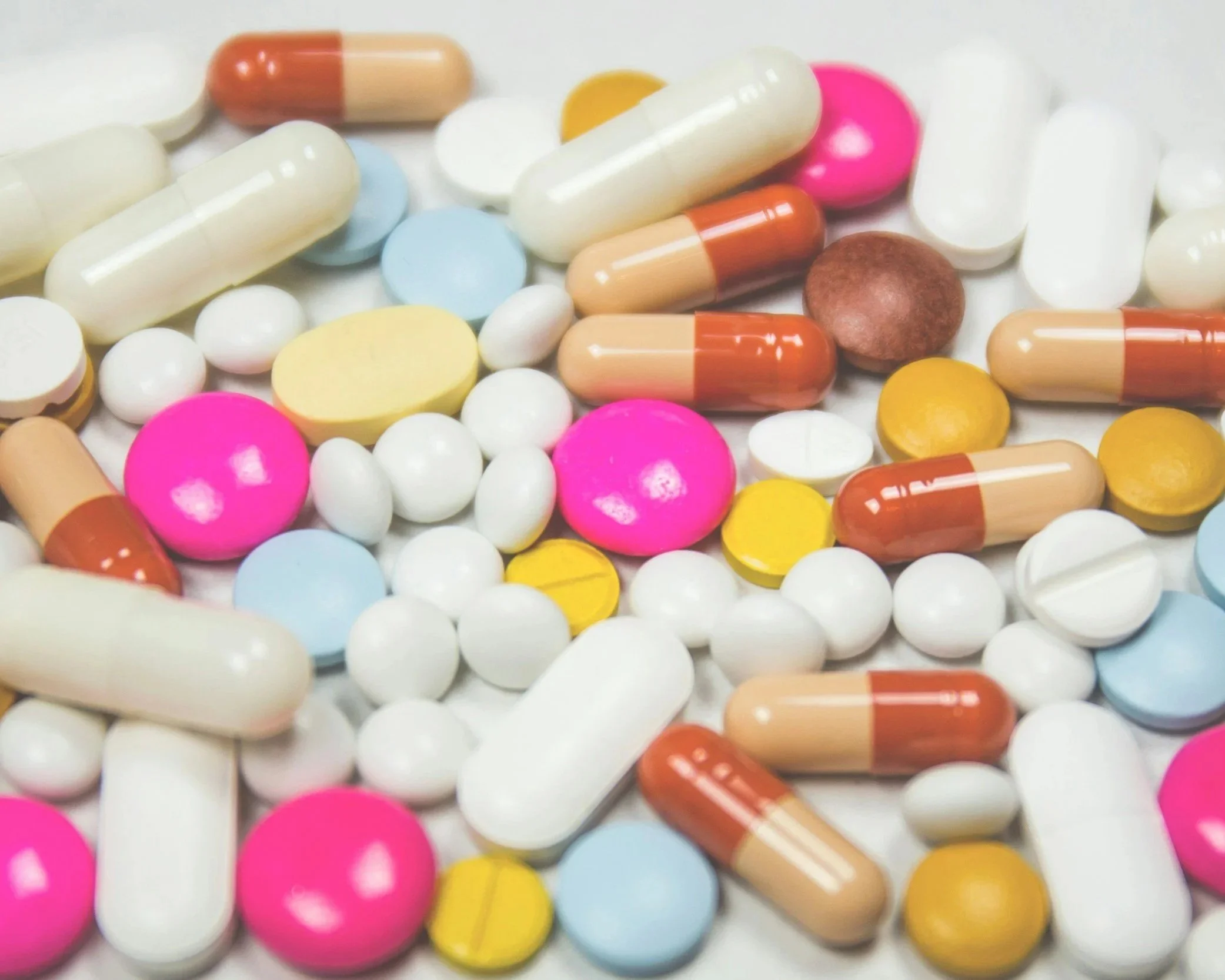 Multicolored pills and capsules scattered on a surface, including pink, white, yellow, blue, and orange in various shapes and sizes.
