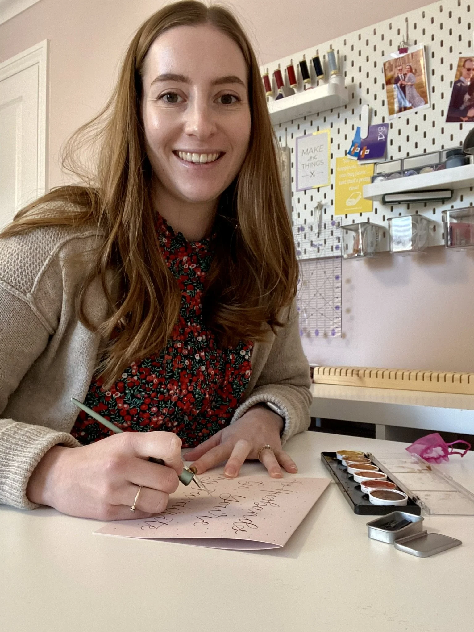 A woman sitting at a desk, smiling, writing a card with a pen, with watercolor paints and a tin case on the table.