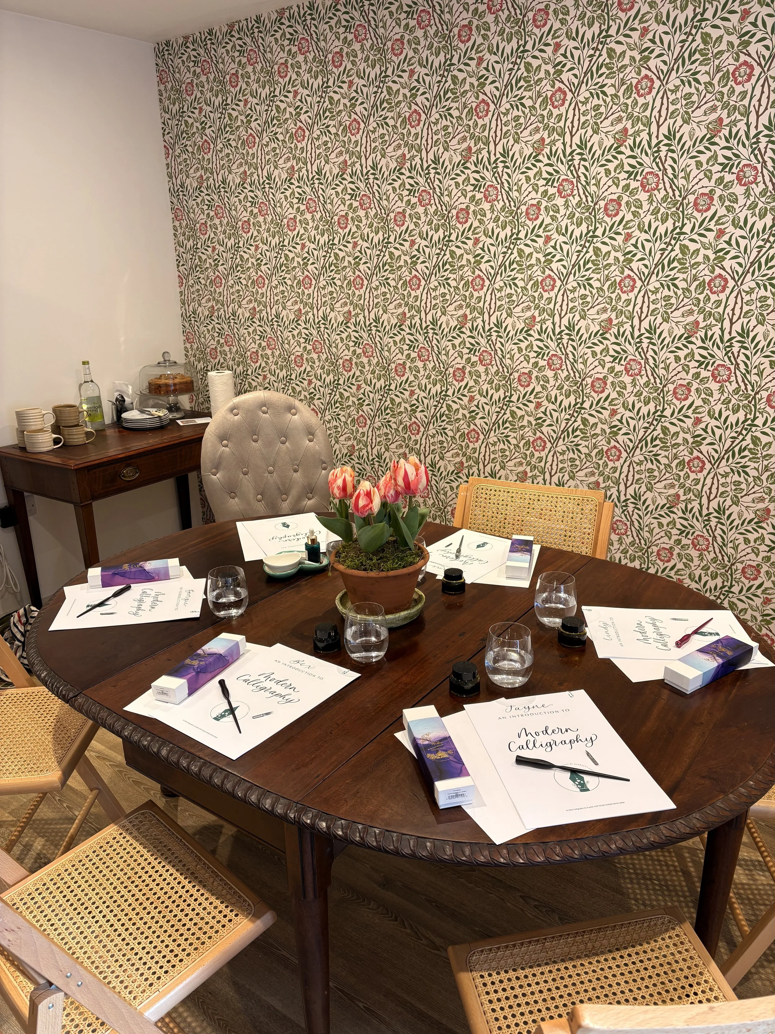 A round wooden table set for a calligraphy class with notebooks, pens, water glasses, and small containers, topped with a potted pink tulip arrangement in a bright room with floral wallpaper and additional chairs around the table.