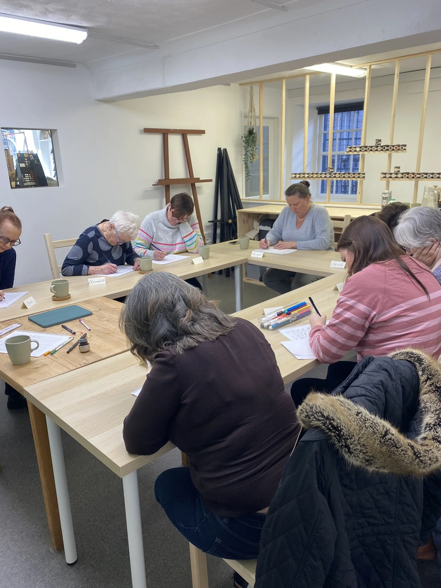 A group of people sitting around a wooden table in a spacious room, taking notes during a meeting or workshop. There are notebooks, pens, and cups on the table.