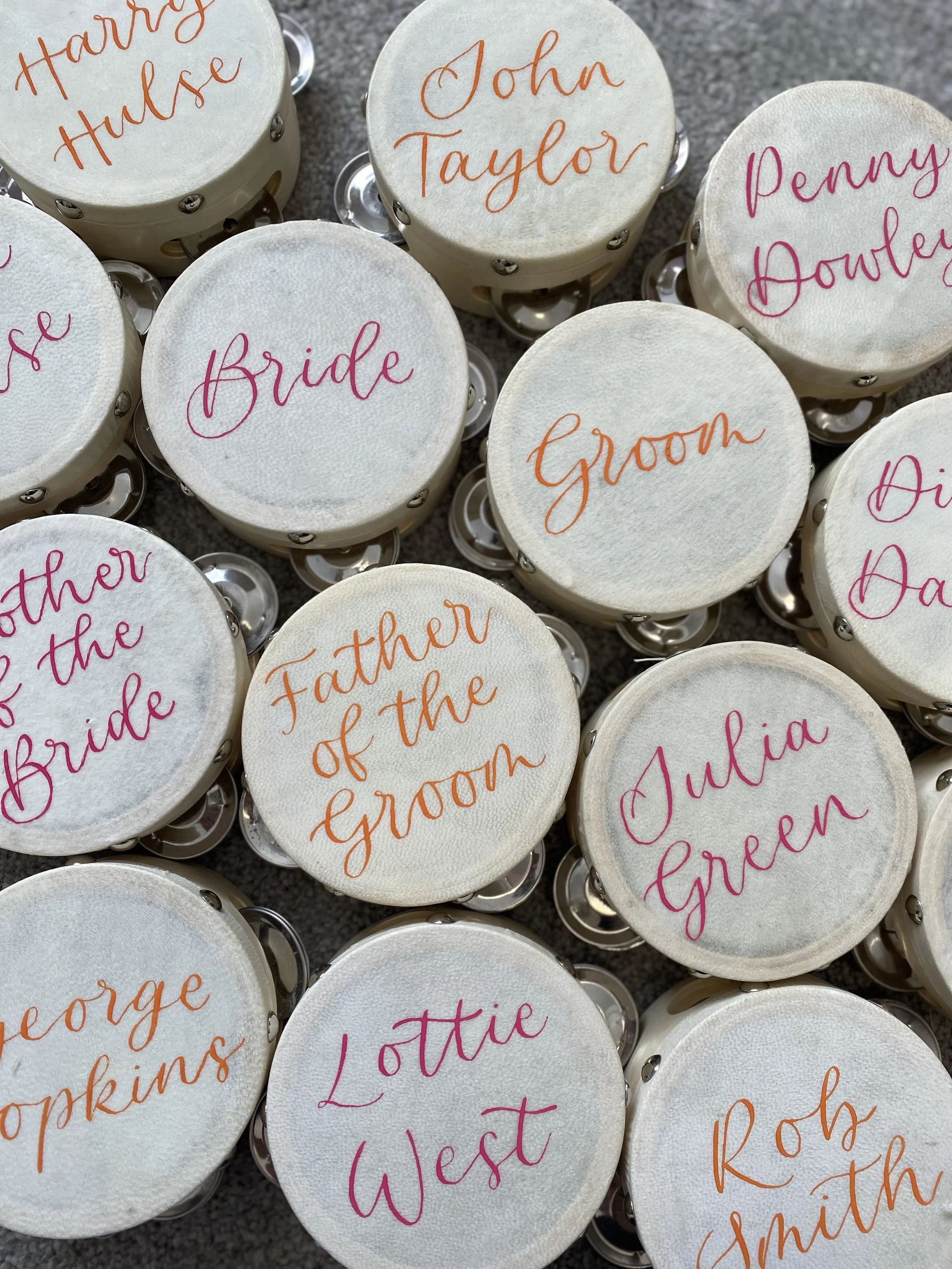 Collection of small round favors with names and titles printed on the tops, for a wedding. Names include Harry Hulse, John Taylor, Penny Downey, Bride, Groom, Father of the Bride, Mother of the Bride, Julia Green, George Hopkins, Lottie West, Rob Smith. The favors are decorated with colorful cursive text.