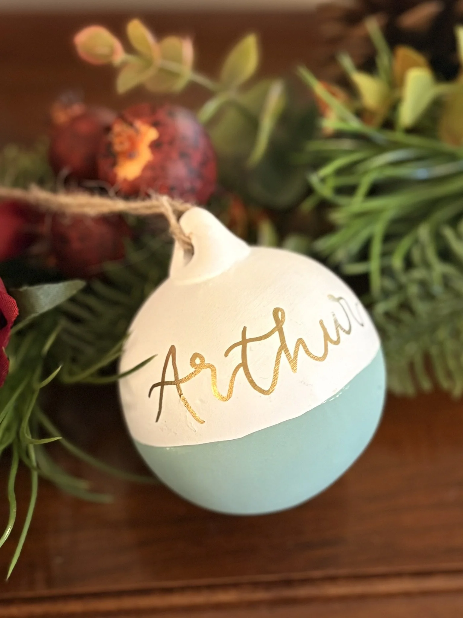 Decorative ornament with the word 'Artful' written in gold script, attached to a string and surrounded by pine branches, pomegranates, and greenery.