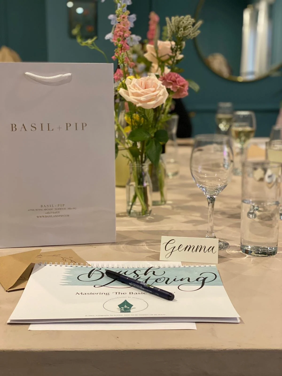 A table set for a flower arranging workshop with a white folder labeled 'Basil + Pip,' a floral centerpiece with pink and white flowers, a name card labeled 'Gemma,' an open booklet titled 'Brush & Mastering,' a black marker, and glasses of water.