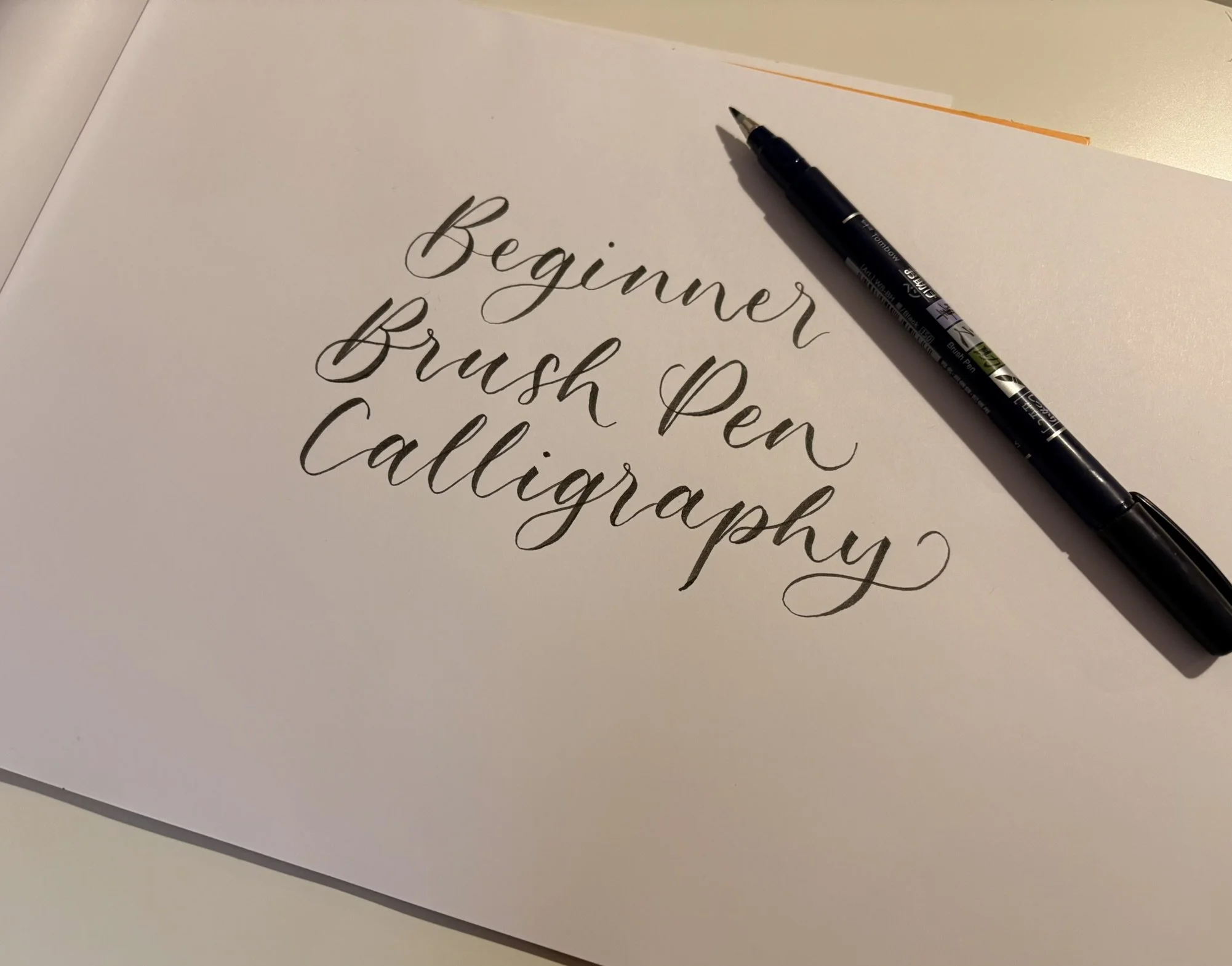 A sheet of paper with the phrase 'Beginner Brush Pen Calligraphy' written in elegant black cursive. A black brush pen rests on the paper at an angle.