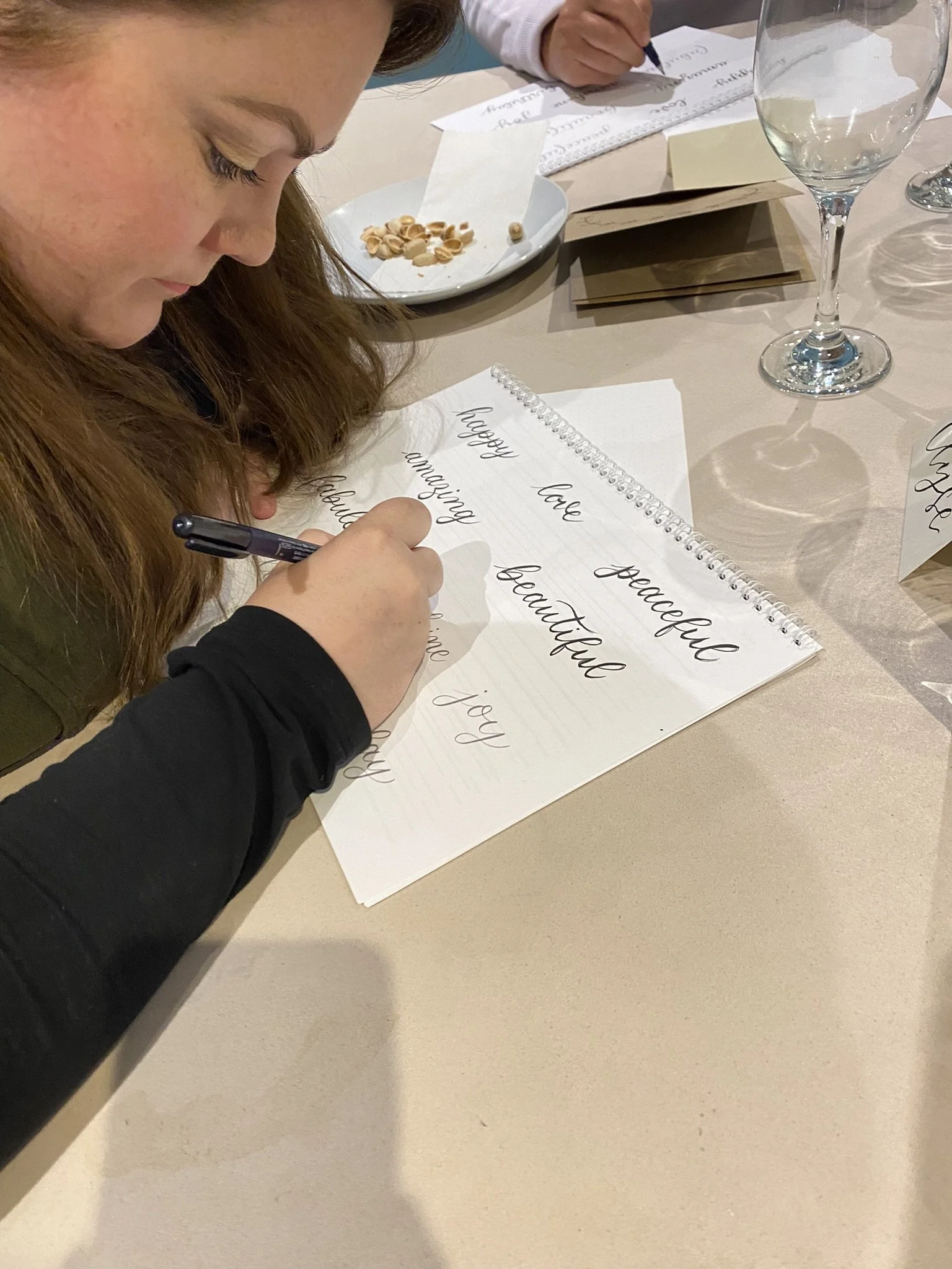 A woman writing calligraphy words like 'love', 'peaceful', 'beautiful', and 'joy' on a white paper with a black pen at a table, surrounded by glassware, paper, and a plate with snack foods.