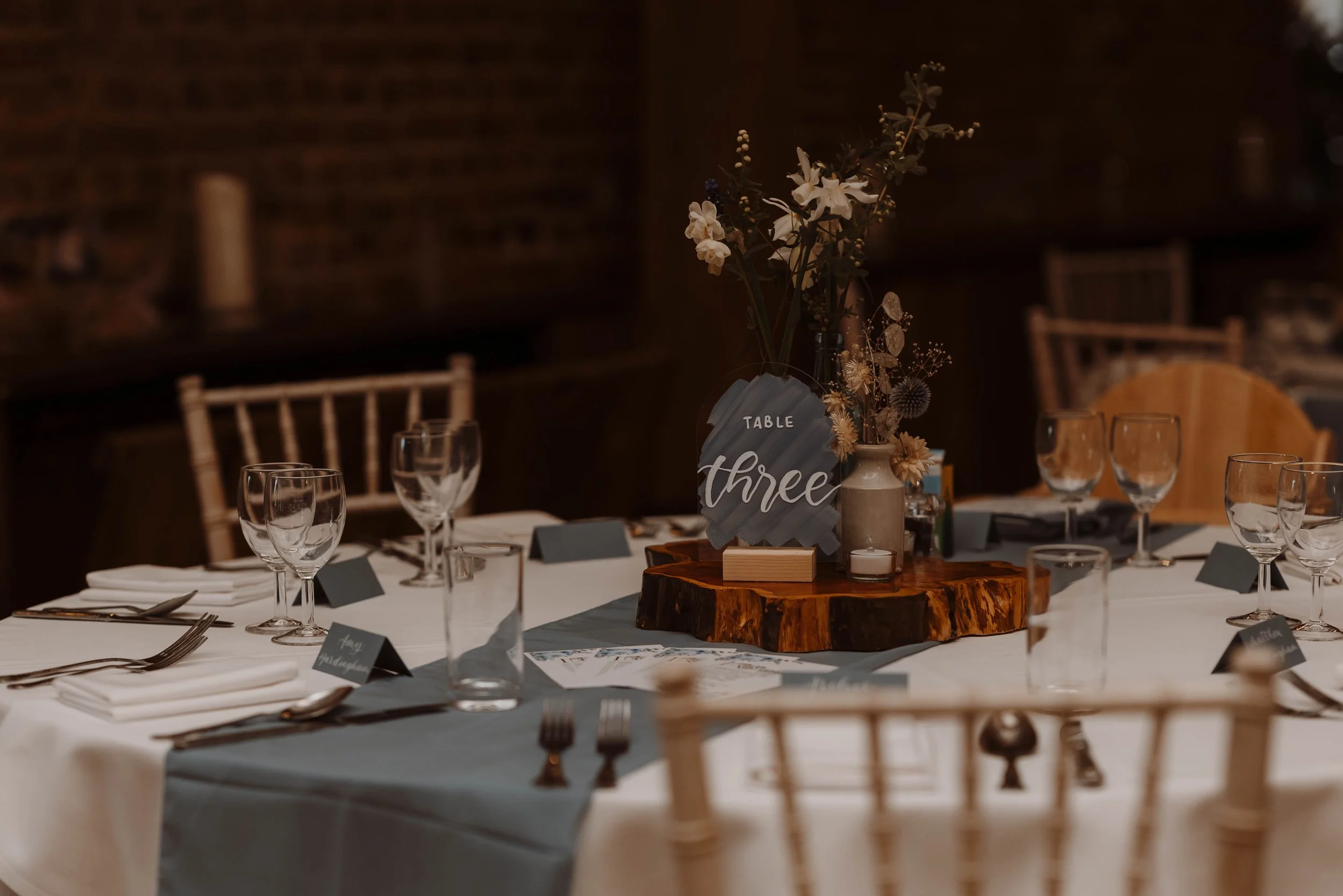 Elegant banquet table set for a formal event with glassware, cutlery, and a centerpiece with flowers on a wooden slab. A sign indicates table three.