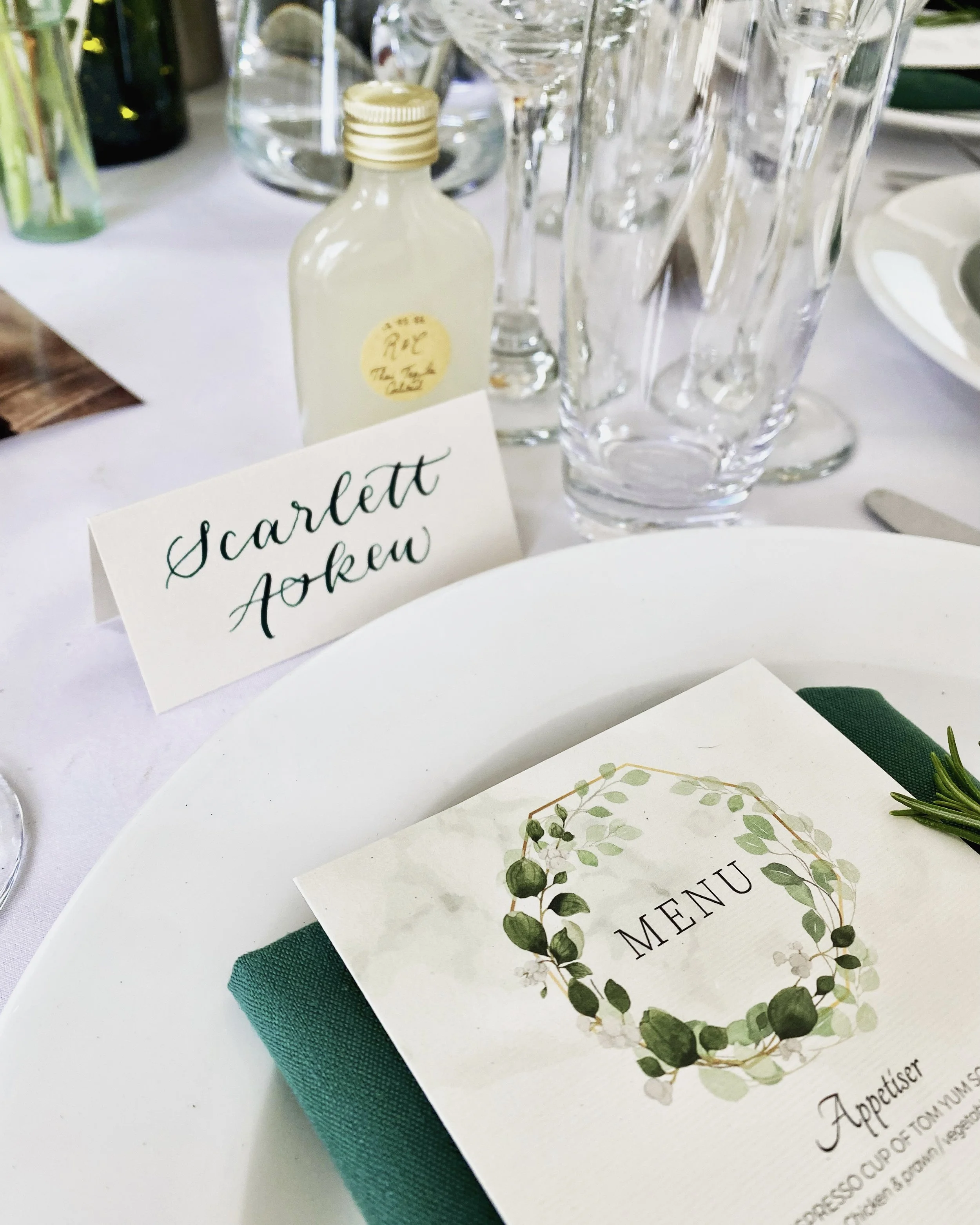 Table setting at a wedding or formal event featuring a menu card with green and white design, a name card reading "Scarlett Jow" in cursive, clear glassware, and a small bottle of what appears to be cream or lotion, on a white tablecloth.