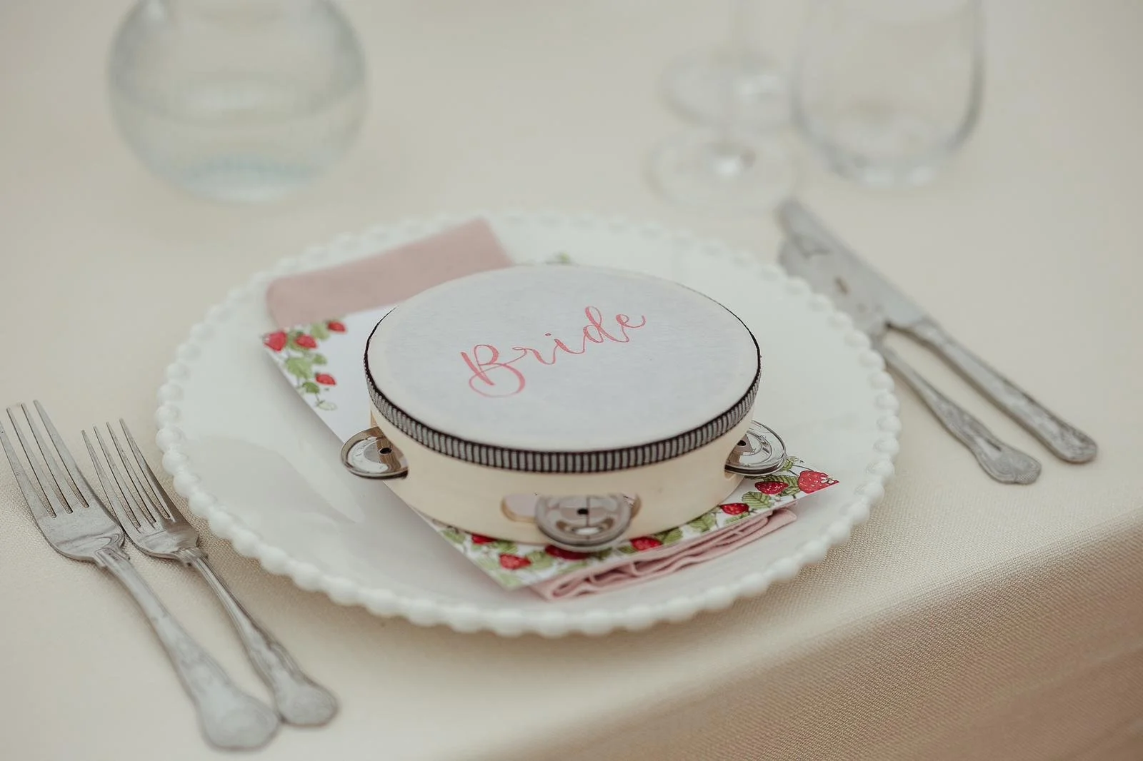 Wedding table setting with a white plate, silverware, a glass of water, and a small white box labeled 'bride' on top of a cloth napkin decorated with strawberries.
