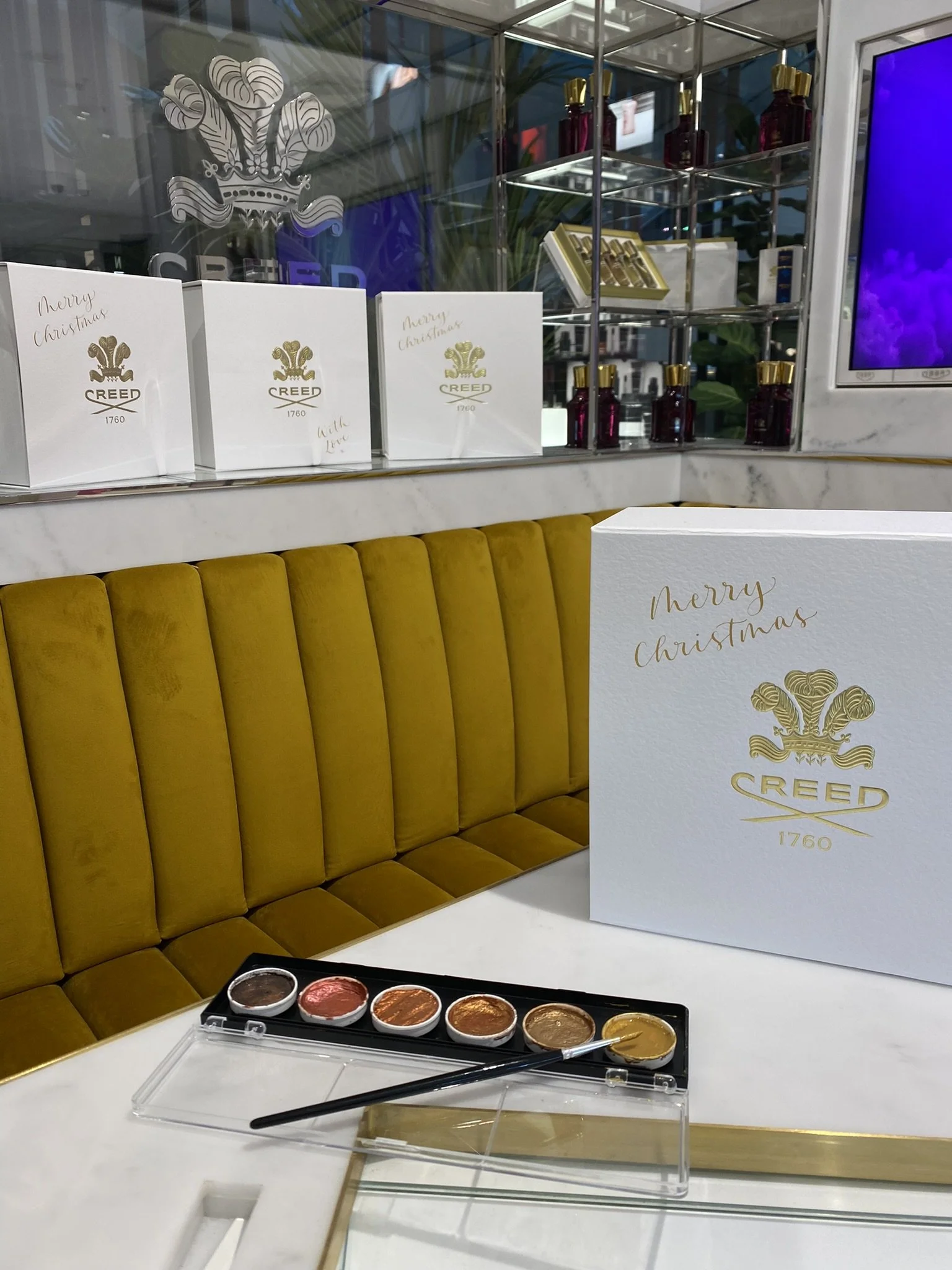 Christmas greeting card from REED 1760, with a palette of paint or makeup swatches in black, pink, red, orange, gold, and yellow, and a brush on a white table in a luxurious setting with a golden-yellow cushioned bench and decorative bottles and boxes on shelves.