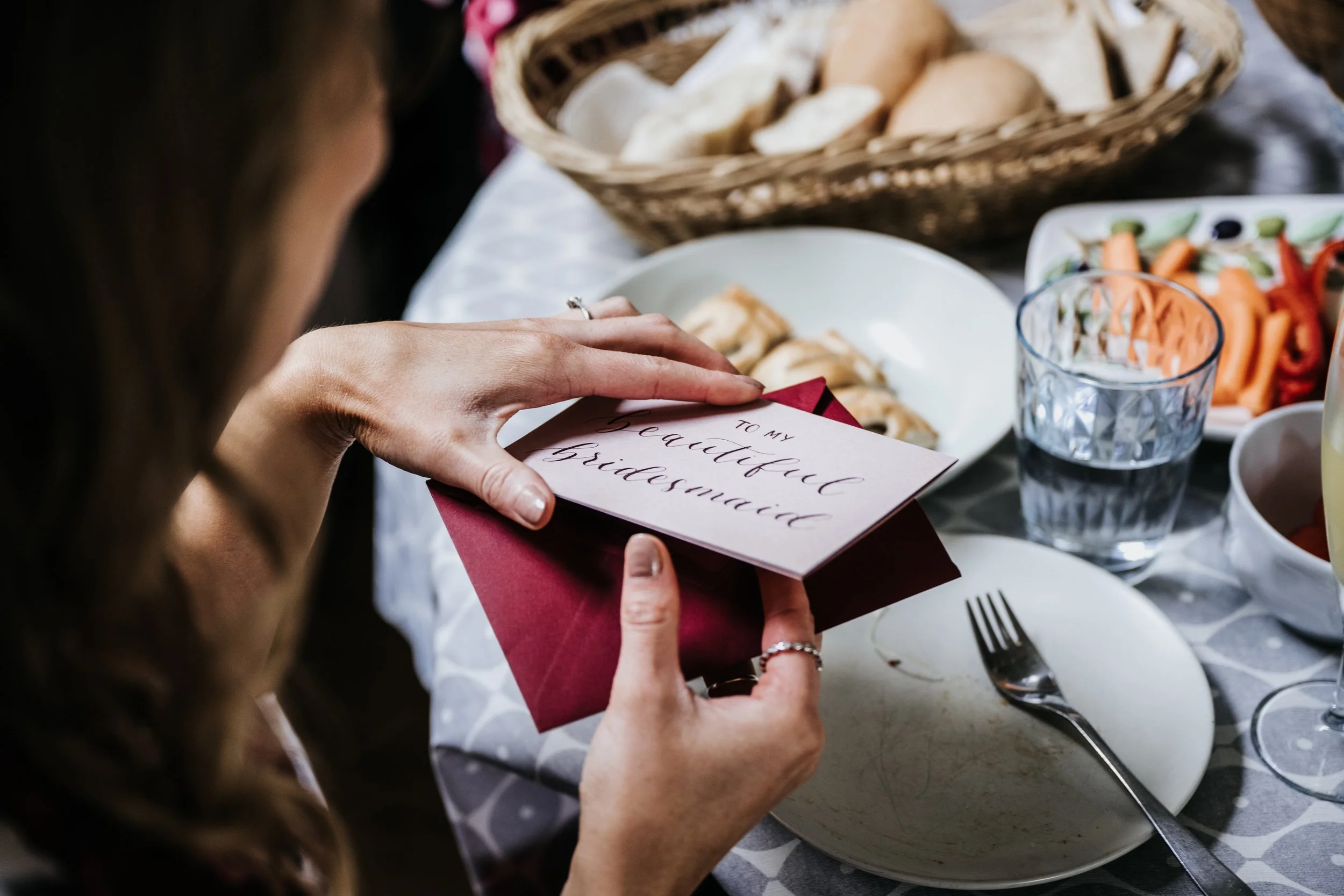 A woman opening a greeting card at a table filled with food and drinks, with a note that reads 'TO MY beautiful bridesmaid'.