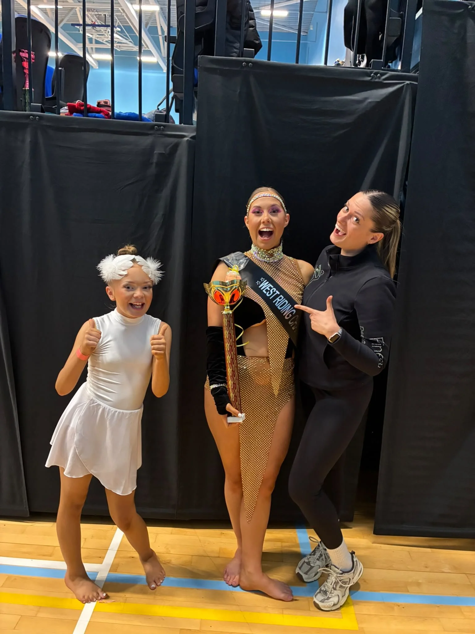 Three smiling performers pose in a gym; the center performer holds a trophy and wears a West Riding sash, while the others give thumbs-up and point toward her.