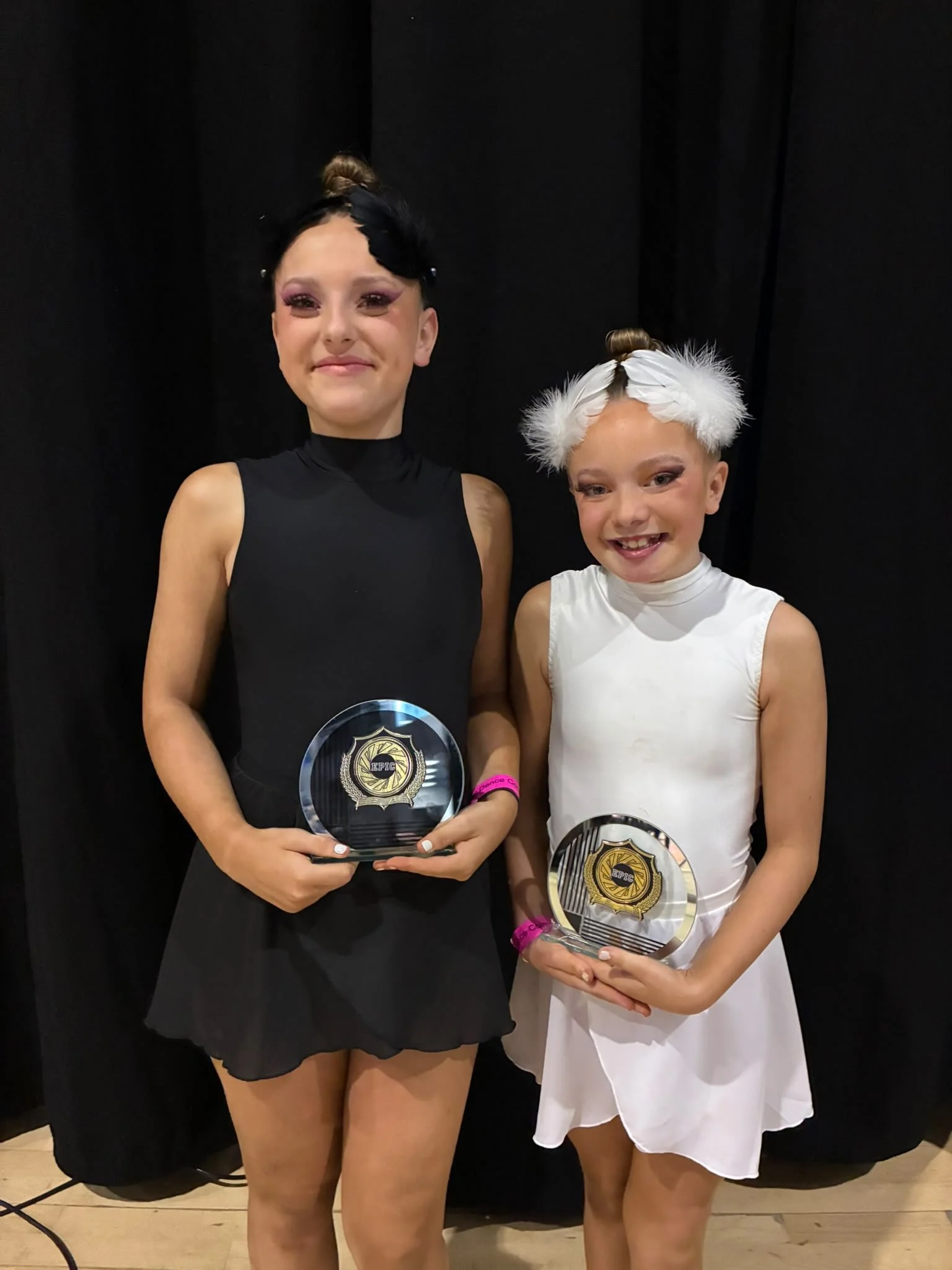 Two smiling young dancers in black and white costumes hold EPIC award plaques in front of a black backdrop.