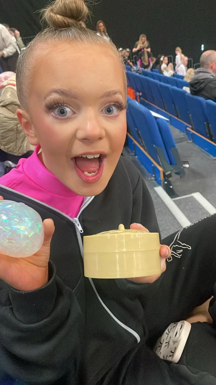 Excited young girl with a top bun holding a yellow snack container and iridescent toy in blue theater seats
