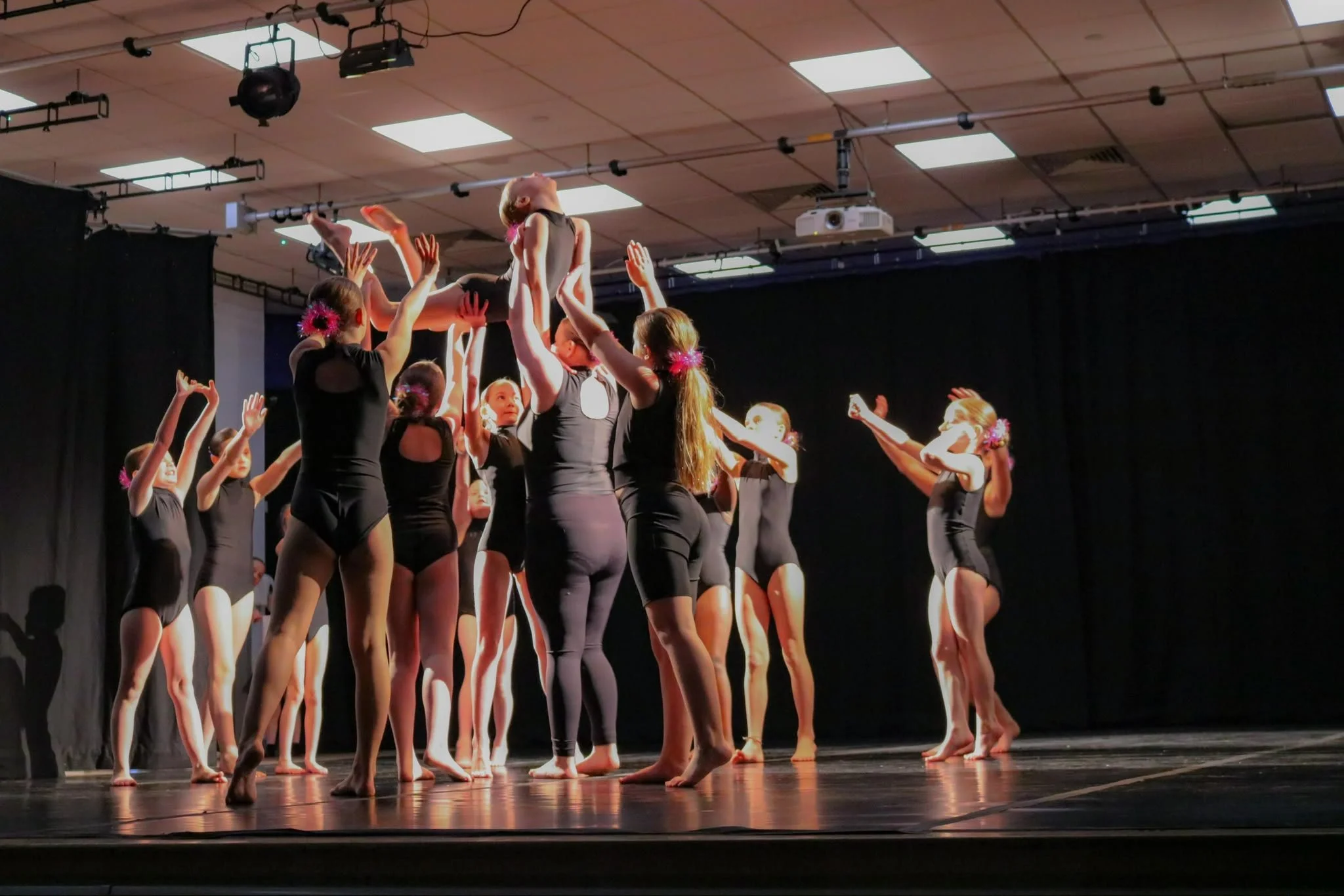 Young dancers in black costumes perform a contemporary dance routine onstage, lifting one dancer under warm theater lights.