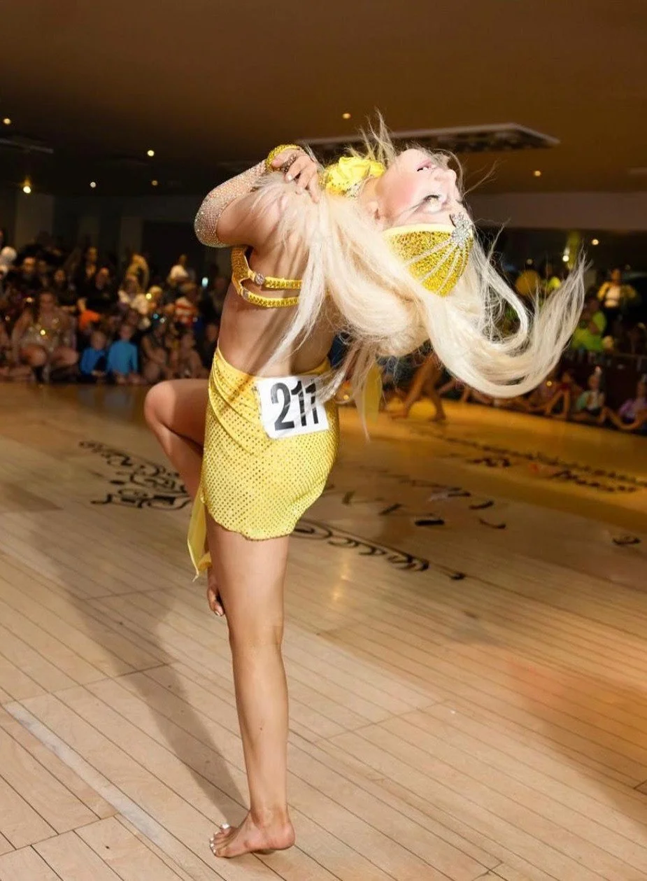 Dancer in a yellow costume and face mask performing on a ballroom floor before an audience