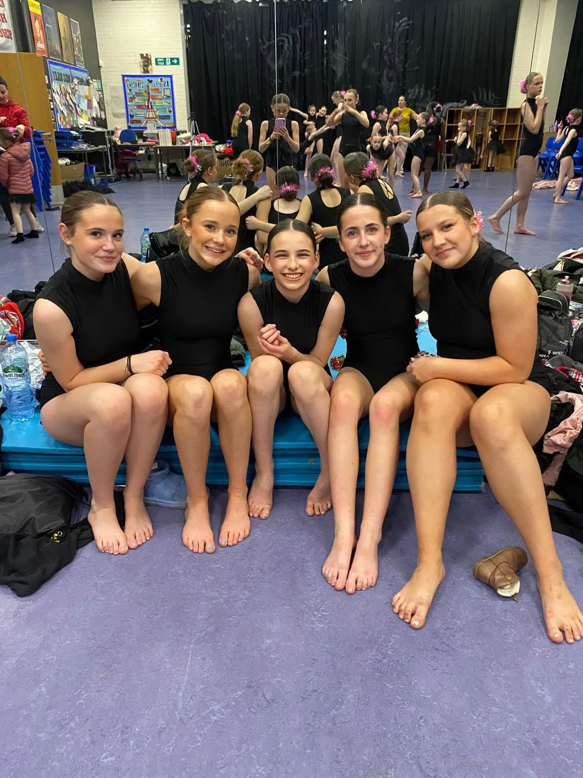 Five smiling young dancers in black outfits pose for a group photo in a mirrored dance studio, with other dancers practicing in the background.