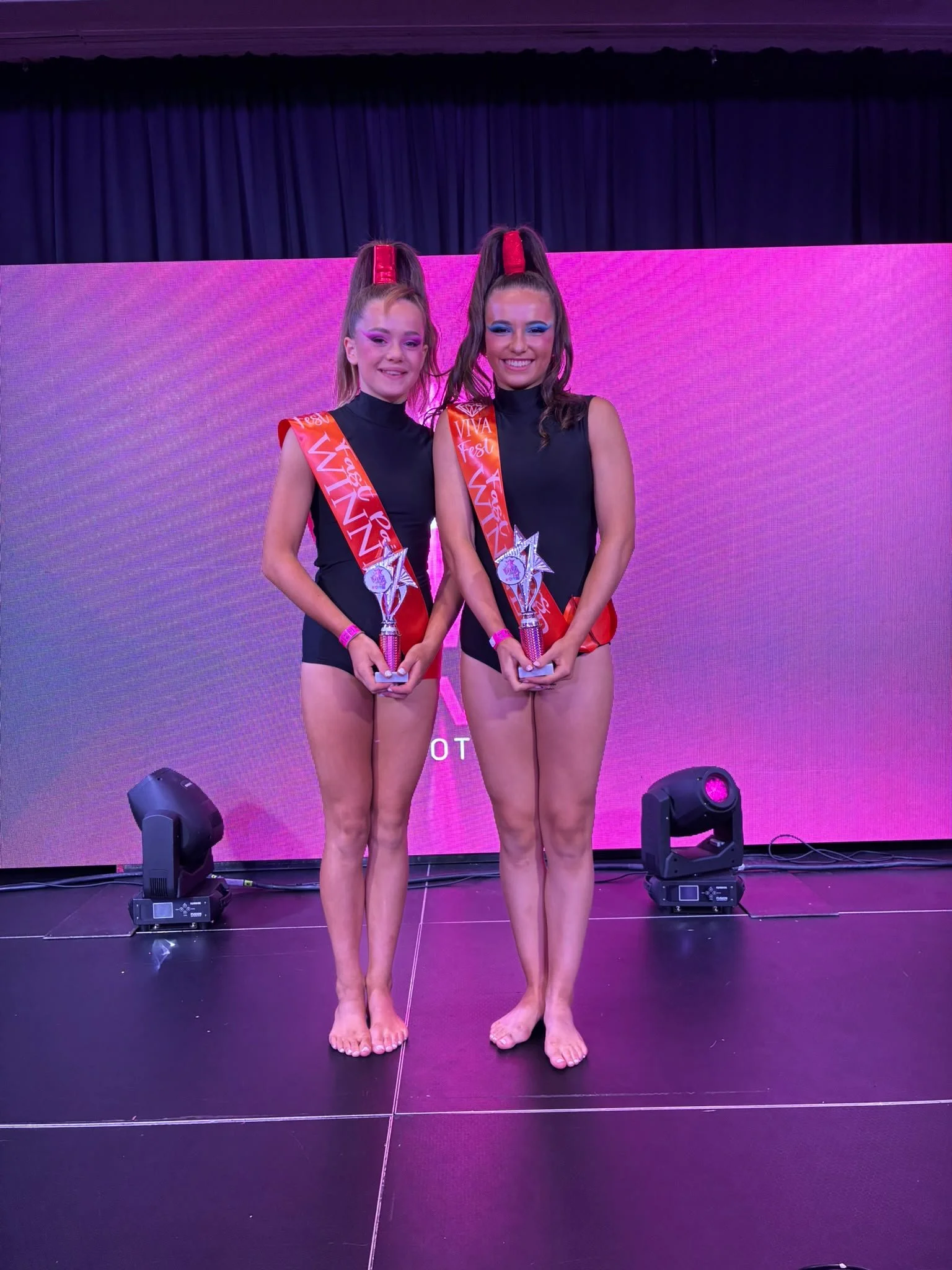 Two smiling women in matching black outfits and red sashes pose onstage holding trophies in front of a pink-lit screen.
