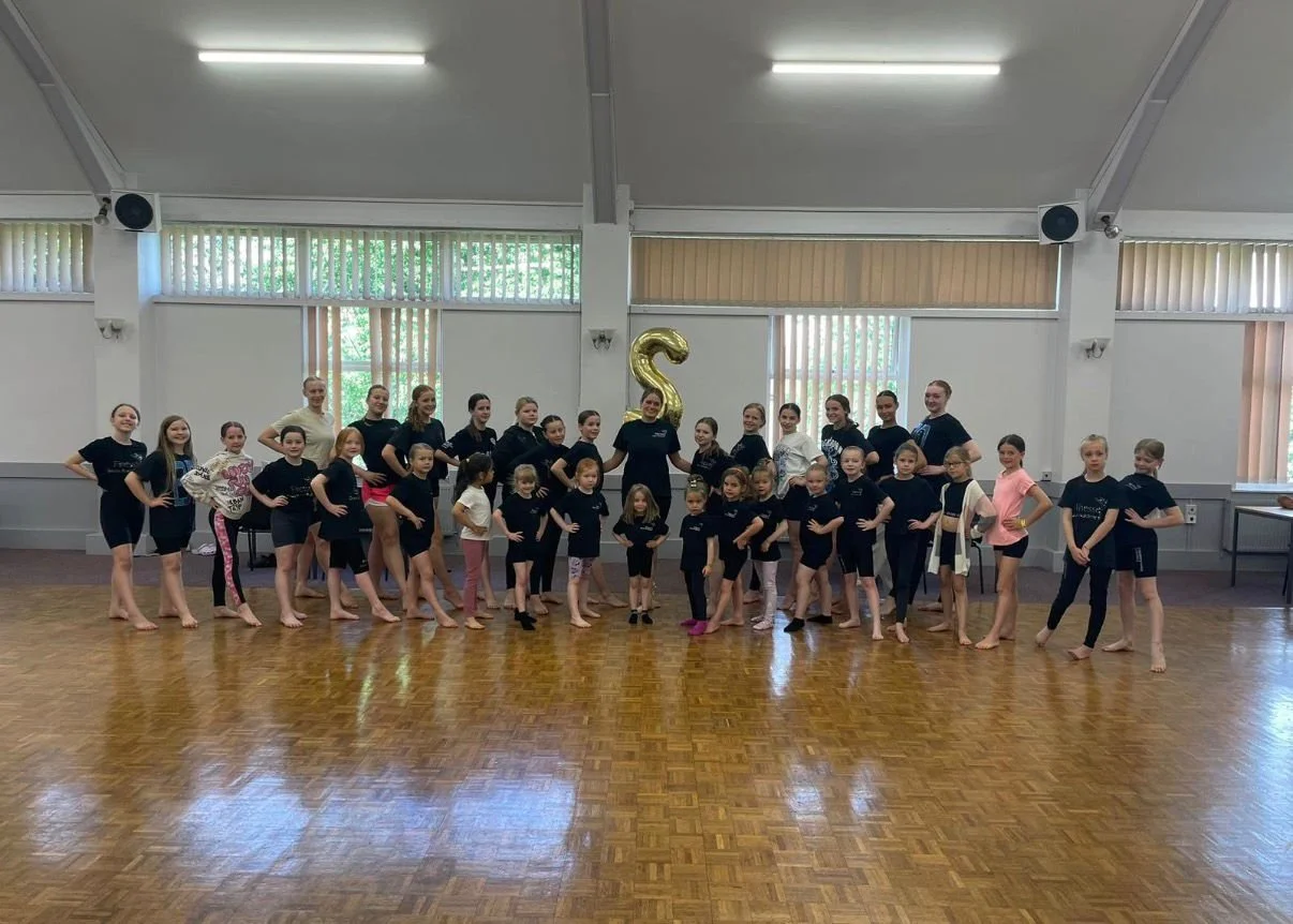 Large group of children and adults posing in a dance studio with a gold number two balloon.