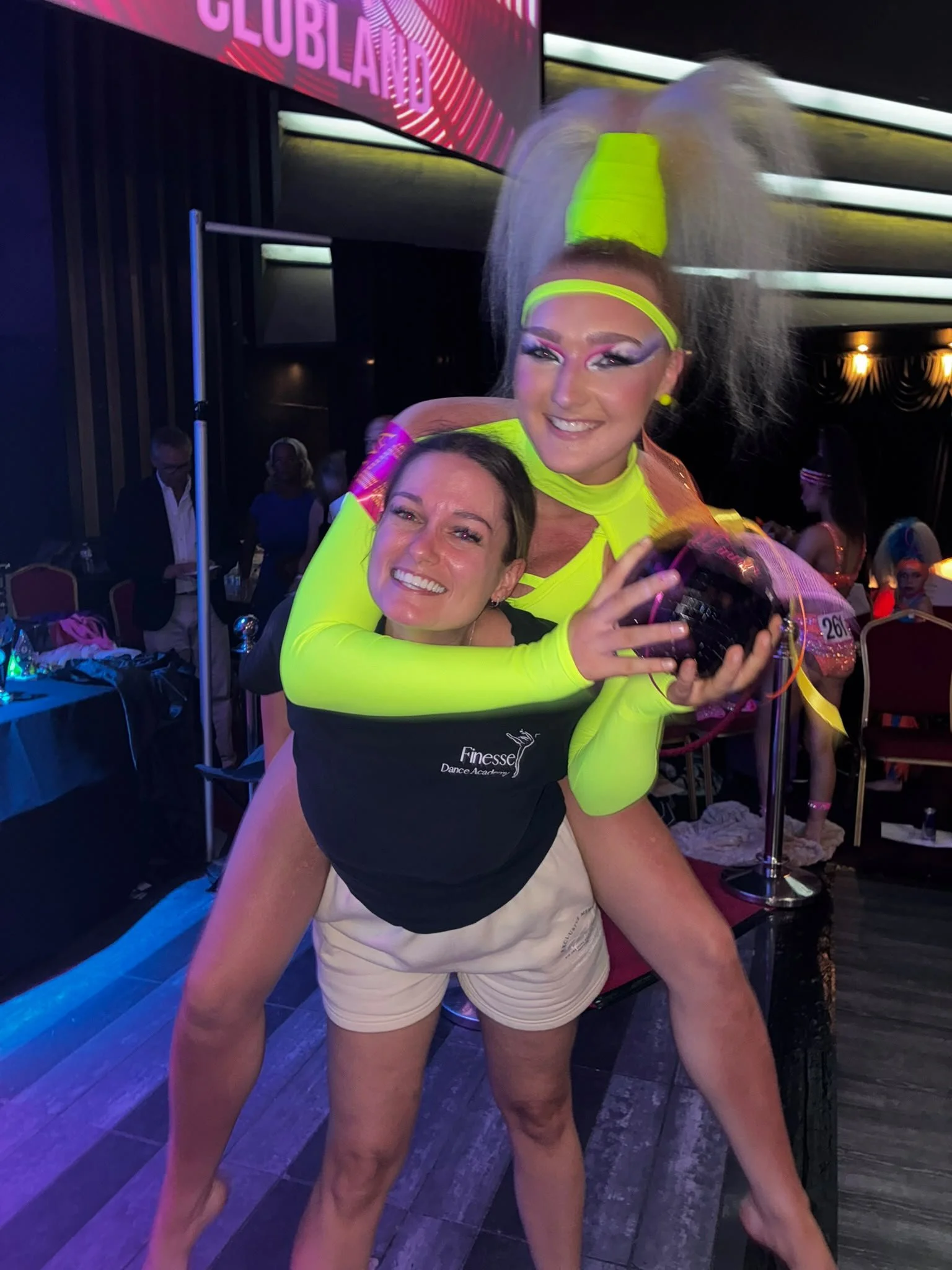 Two smiling women pose at a neon-lit dance event, with one woman in a bright green costume hugging the other from behind.
