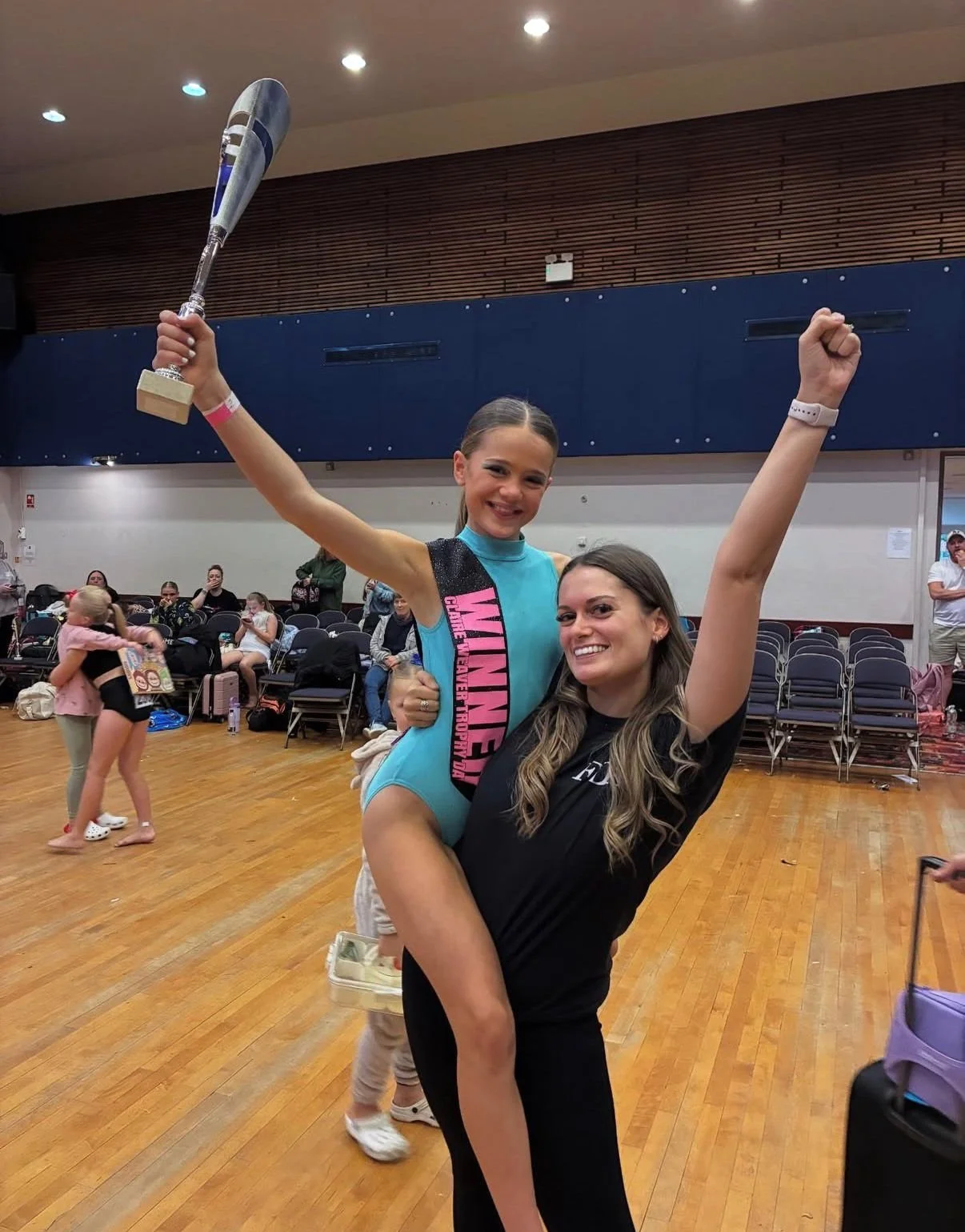 Young gymnast holding a trophy while riding on a smiling woman’s back in a gymnasium