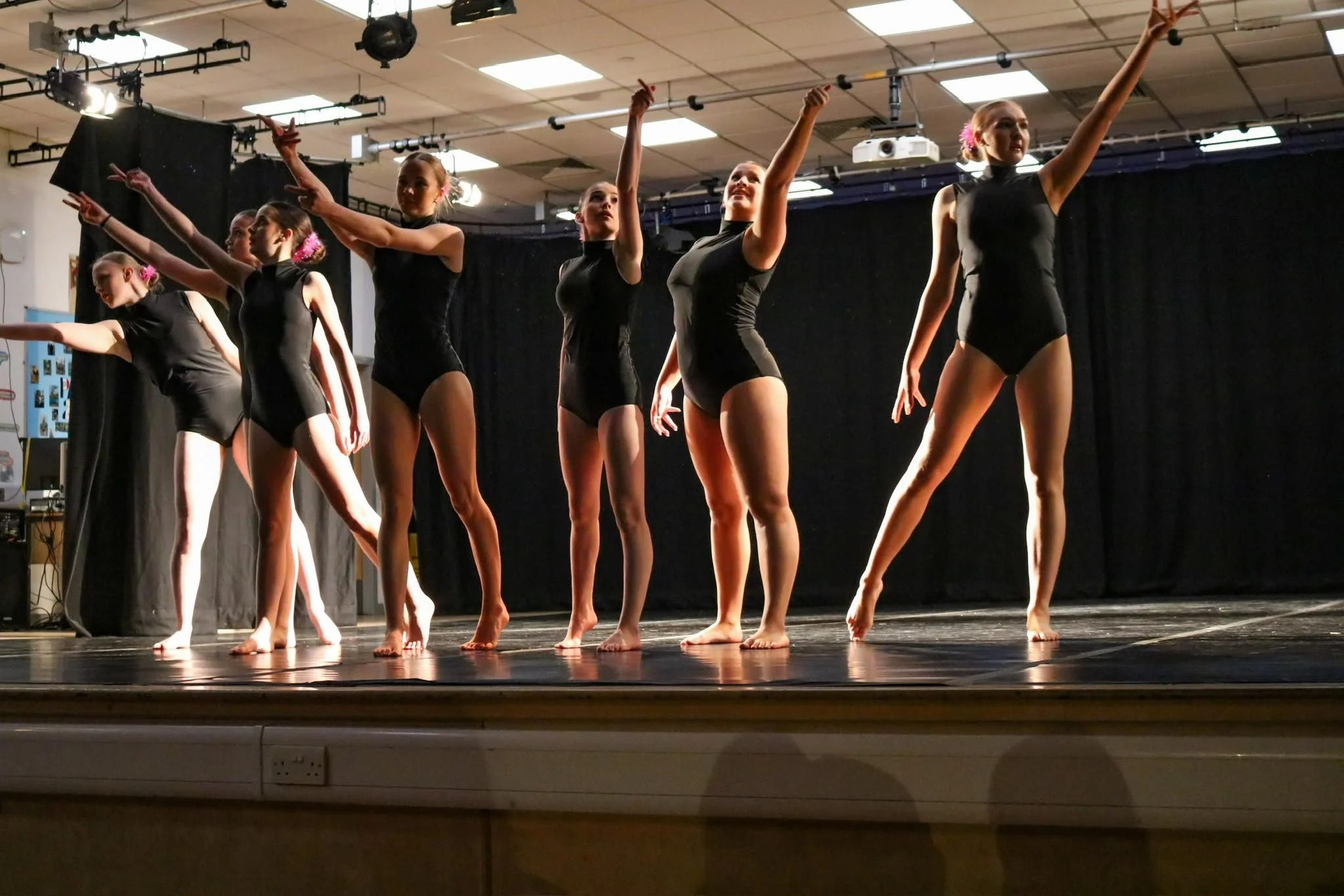 A group of female dancers in black leotards performing on a theater stage under bright lights.