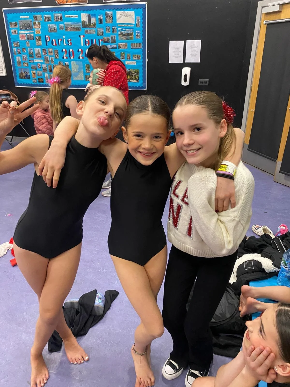 Three young girls pose together in a dance studio, with two wearing black leotards and a Paris Tour 2023 photo board in the background.