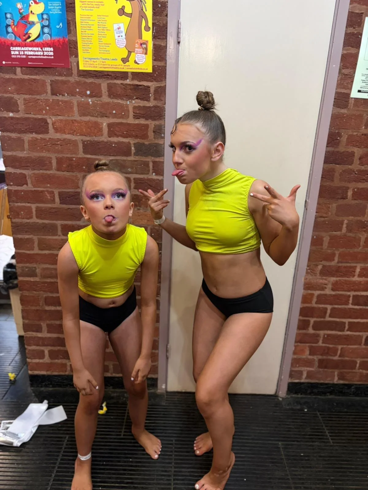 Two female dancers in neon yellow crop tops and black shorts pose playfully in a backstage hallway.