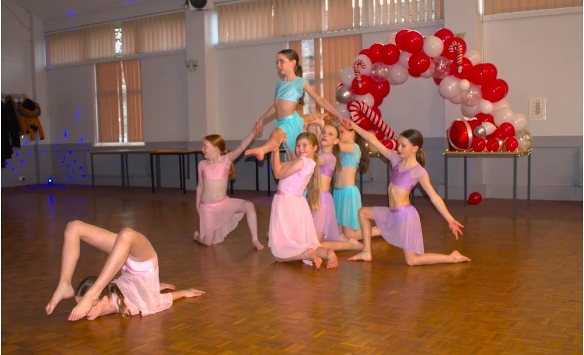 Young dancers in colorful costumes perform a choreographed pose on a studio floor beneath a red-and-white balloon arch.