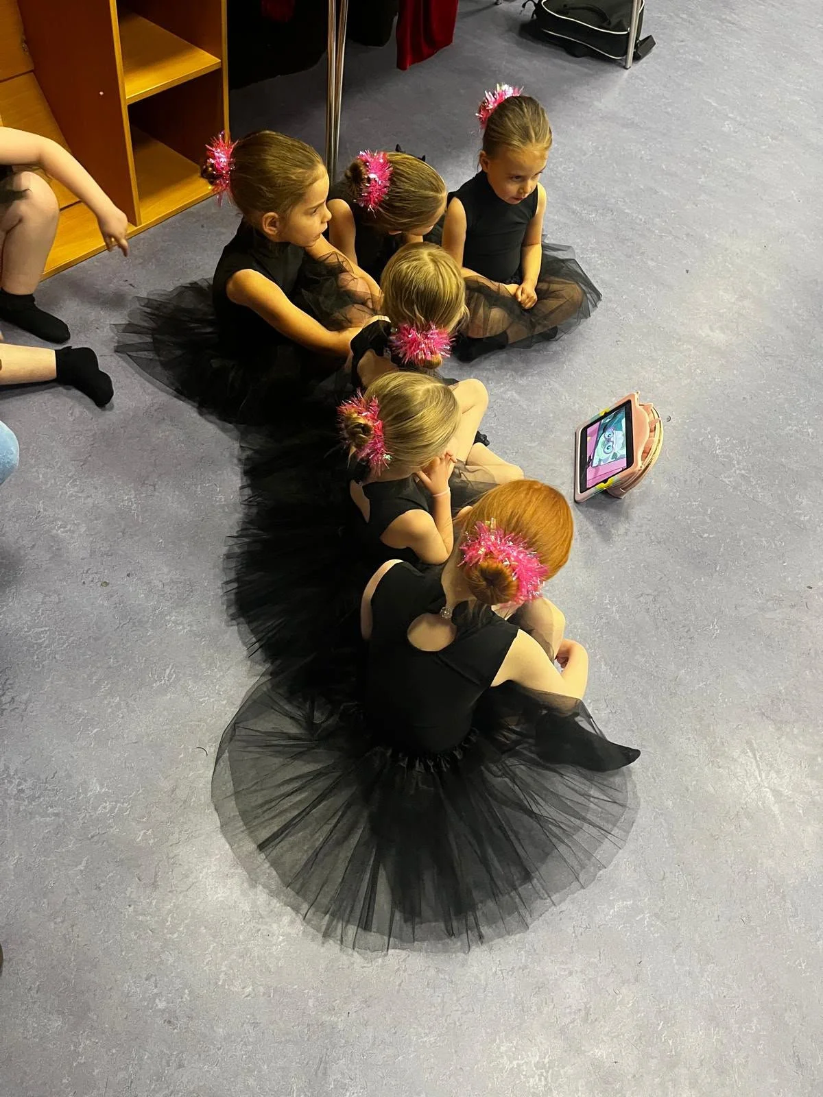 Young ballet dancers in black tutus and pink hair accessories sit together on a studio floor.