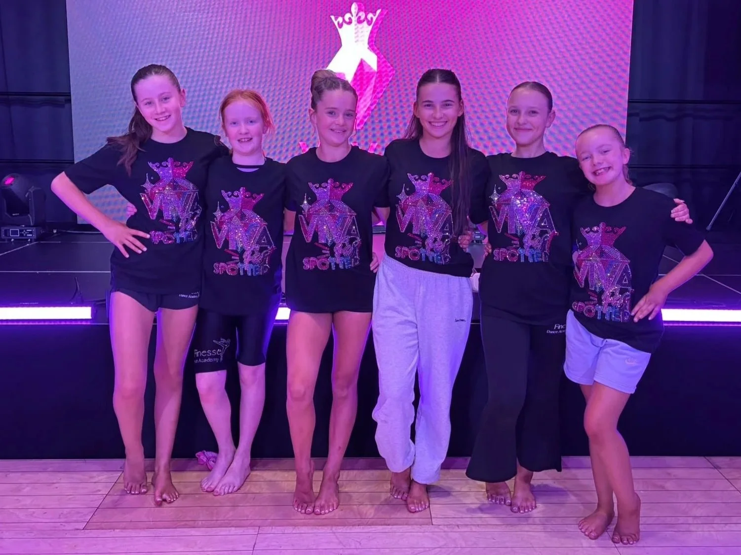 Six smiling young dancers posing onstage in matching black MA dance team shirts under purple lighting.